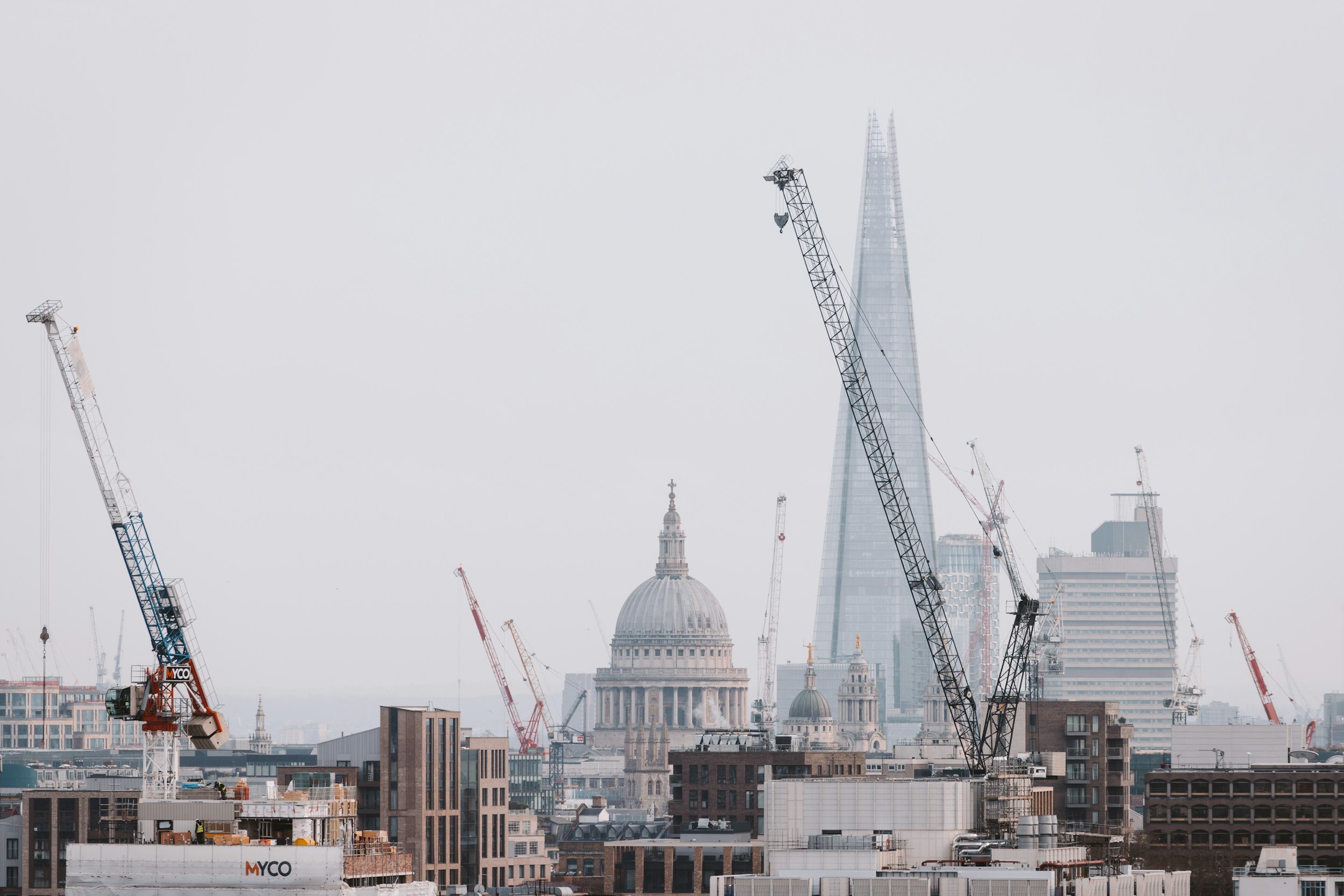 City skyline with multiple construction cranes and the Shard skyscraper in the background under a cloudy sky.