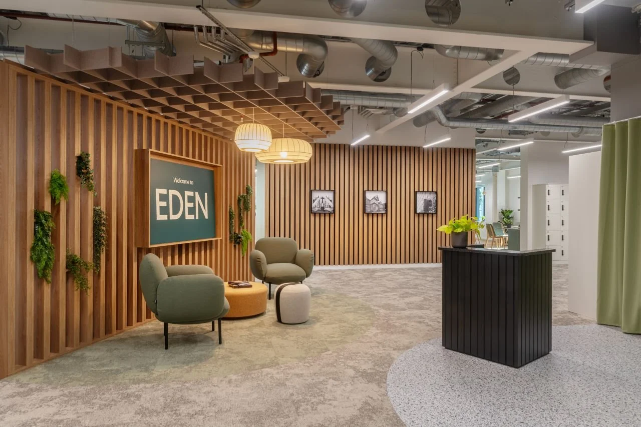 Modern office reception area with a wooden slat wall, two hanging pendant lights, a large sign that reads 'Welcome to EDEN,' two green armchairs, a small ottoman, a black reception desk with a plant, framed black-and-white photos on a wooden partition, and green curtains on the right side.
