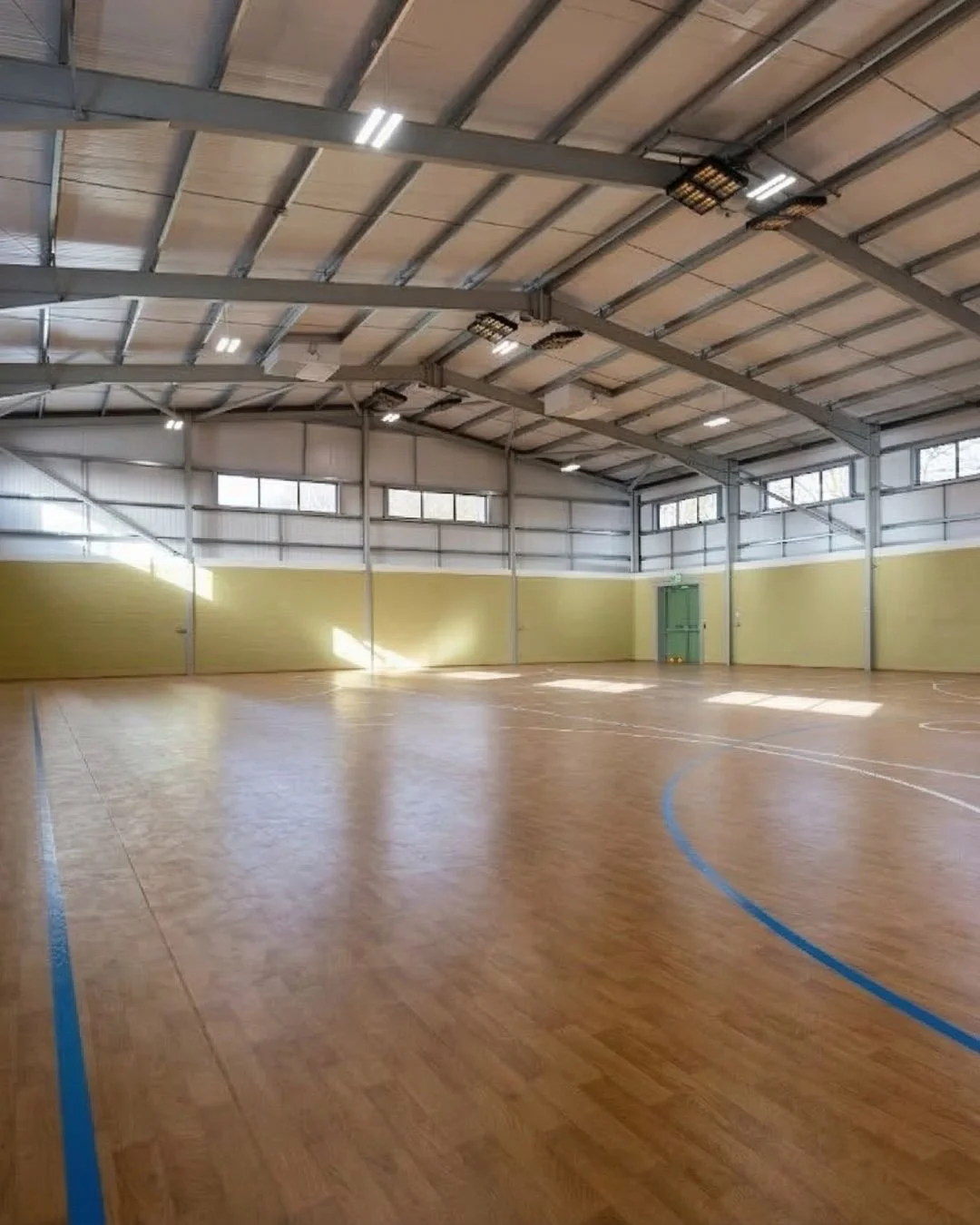 An empty indoor sports gymnasium with a wooden floor, yellow walls, and large windows near the ceiling. Green doors are visible on one side of the room.