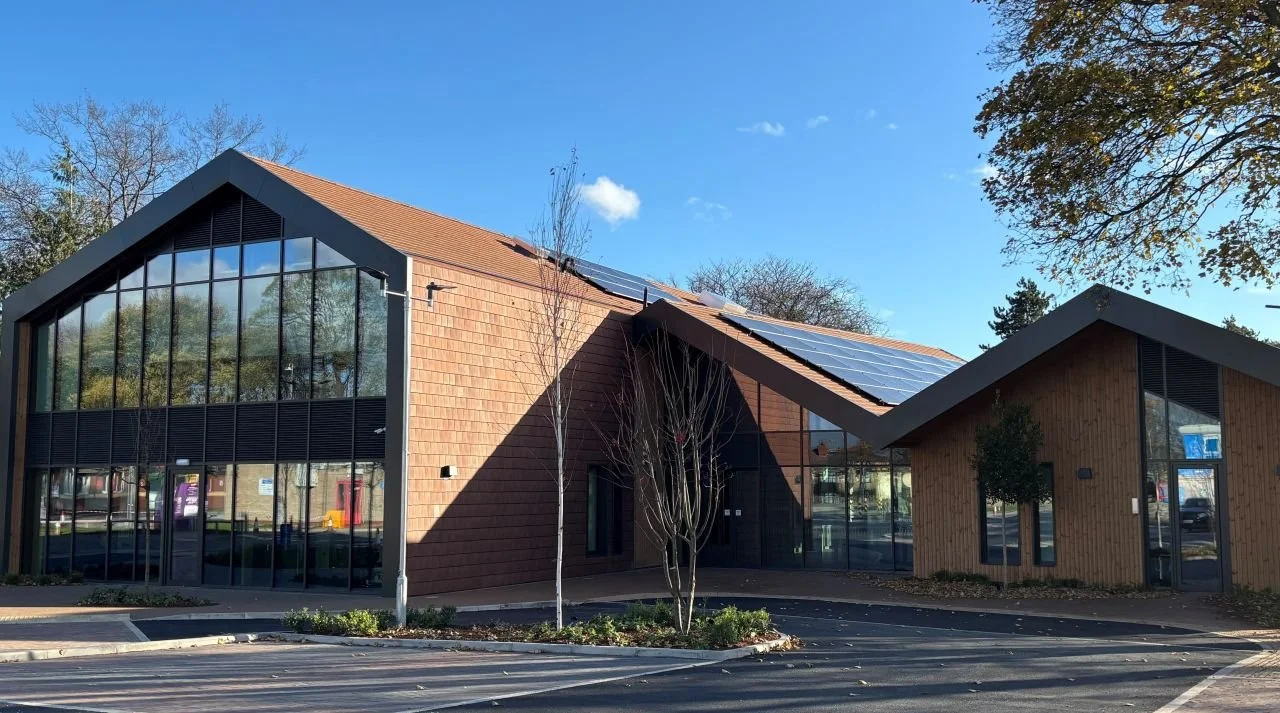 Modern building with a sloped roof, large glass windows, and wooden exterior, situated in a parking lot with leafless trees and a clear blue sky.