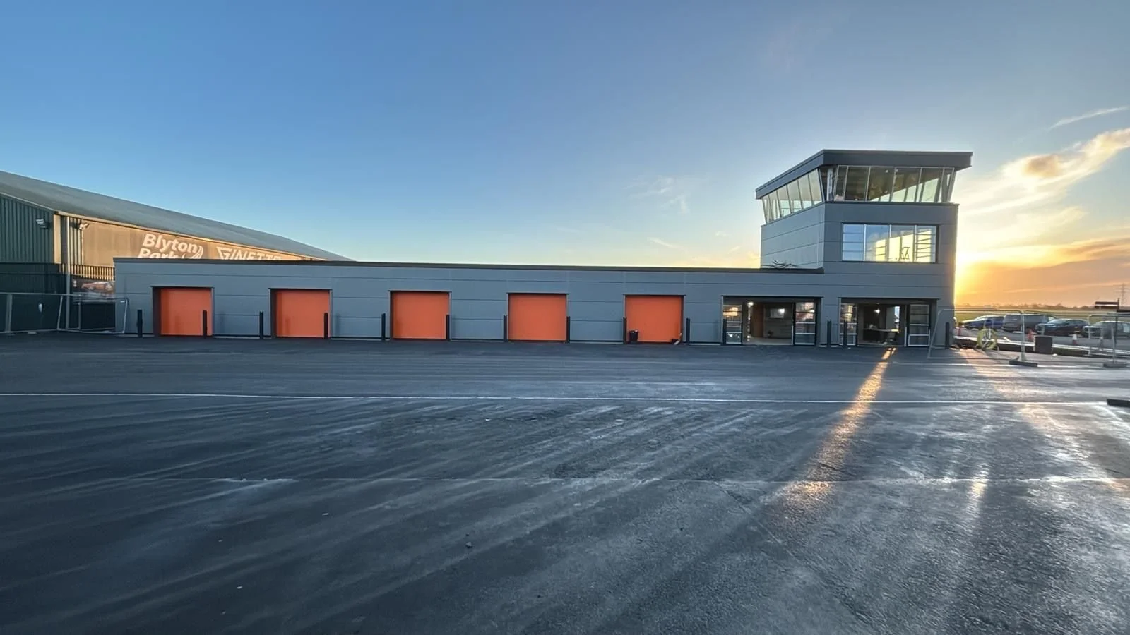 Empty parking lot in front of a modern building with orange garage doors, a glass tower, and sunset sky.