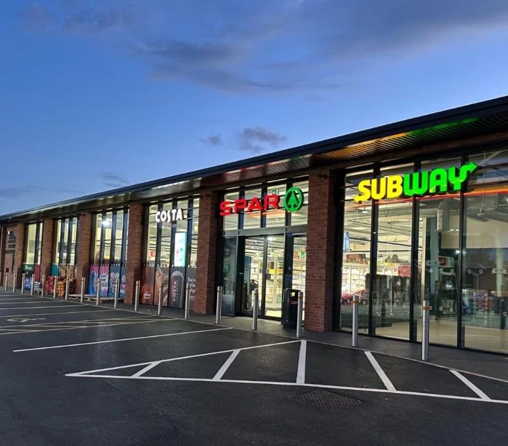 An exterior view of a convenience store shopping strip featuring signs for Costa, Spar, and Subway, with empty parking spaces and a partly cloudy evening sky.