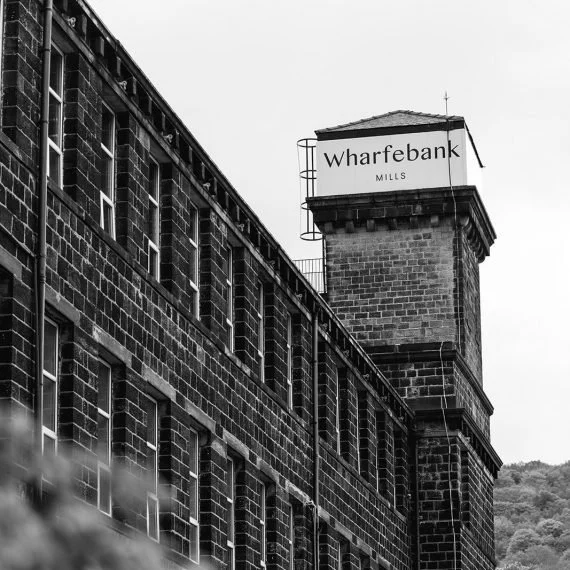 Black and white photograph of an old industrial brick building with a tower that has a sign reading 'Wharfebank Mills'.