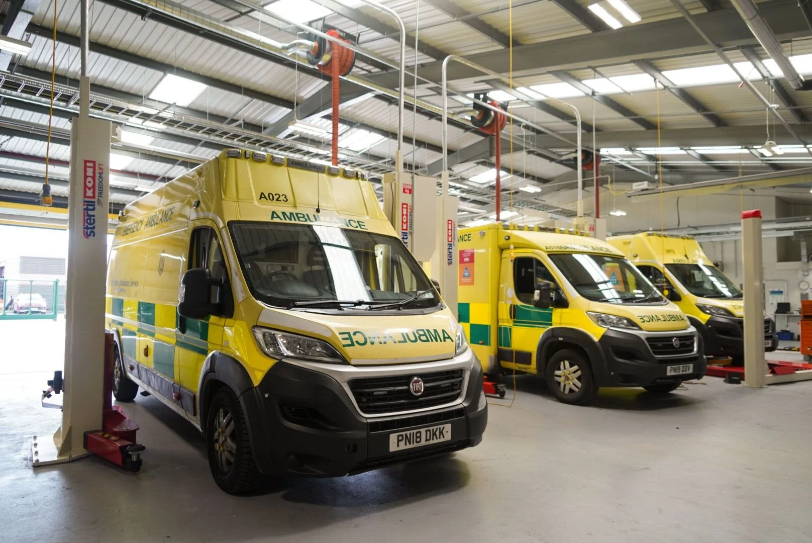 Three yellow ambulances parked inside a vehicle maintenance garage, with various equipment and overhead lighting.