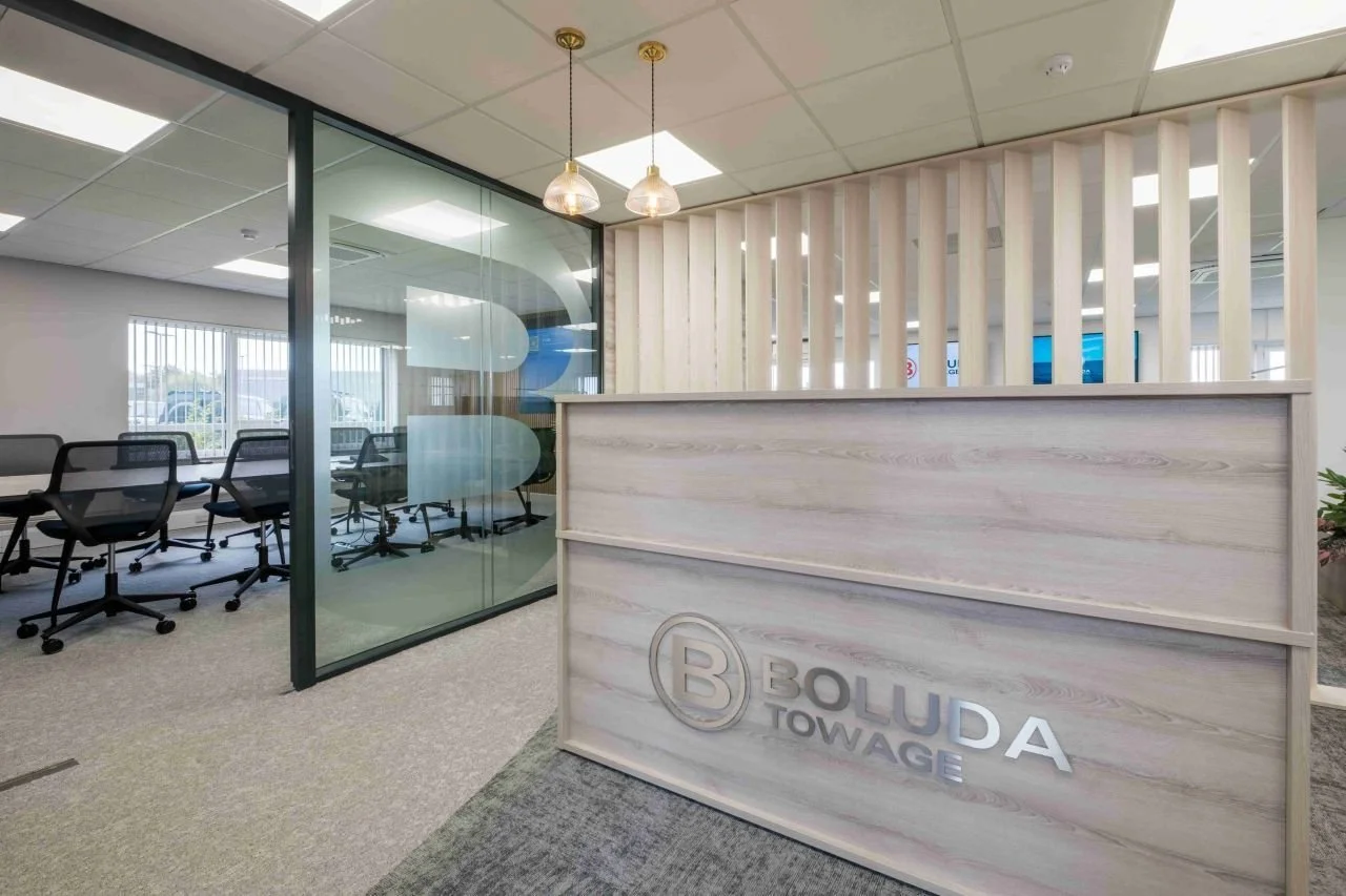 Office reception area with a wooden desk displaying the logo 'BOLUDA TOWAGE', glass wall with office chairs inside meeting room, pendant light fixtures, and a window with blinds in the background.