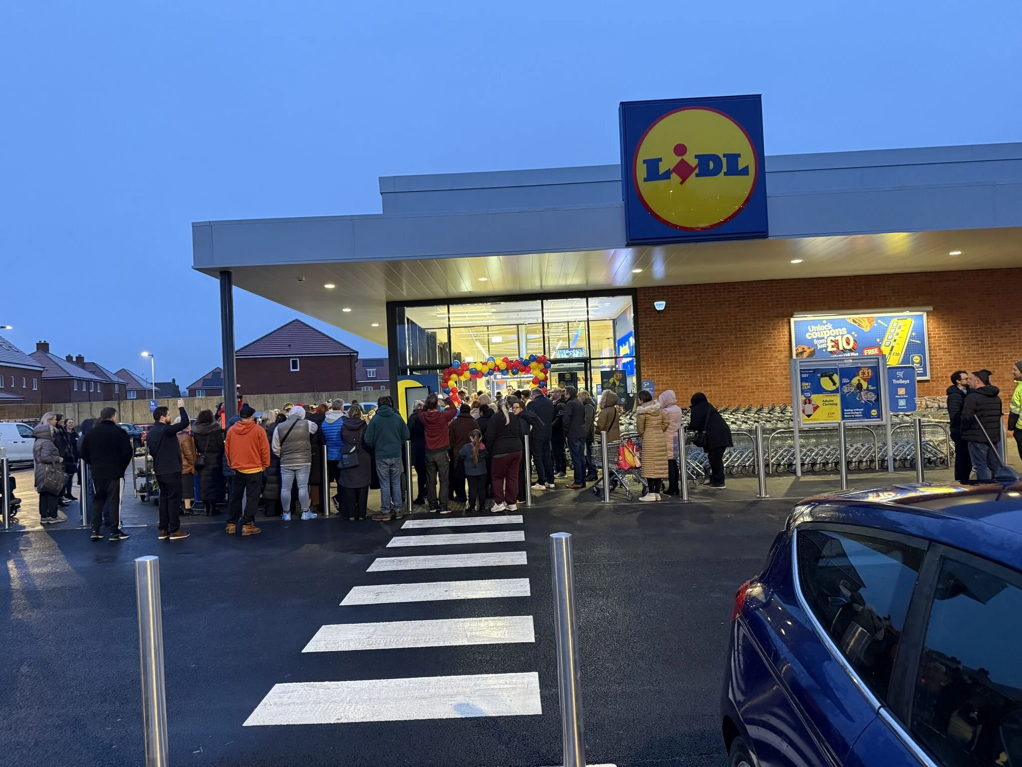 Large crowd of people lined up outside a Lidl grocery store during evening hours with a bright sign and balloons at the entrance.
