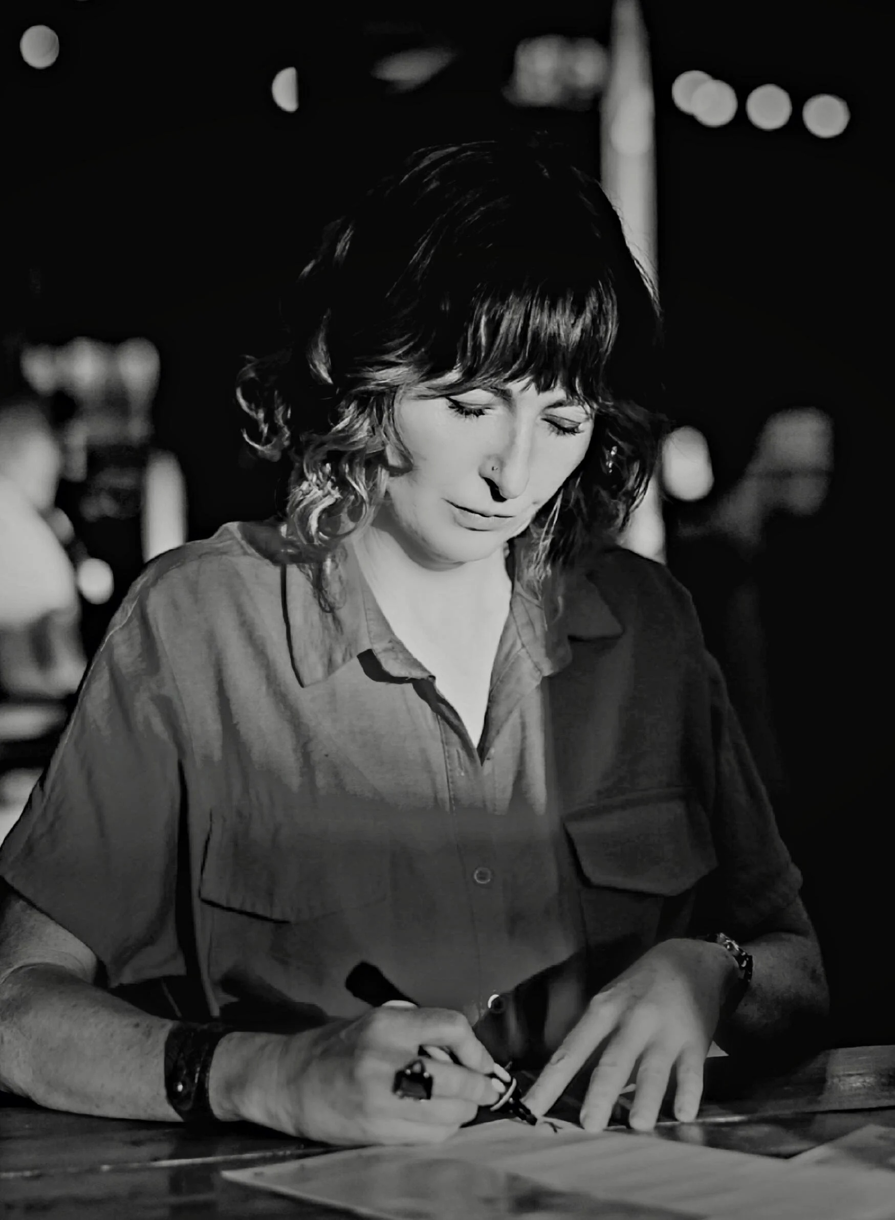Black and white photo of a woman with wavy hair, wearing a button-up shirt, writing on paper with a pen, sitting at a desk in a dimly lit setting.