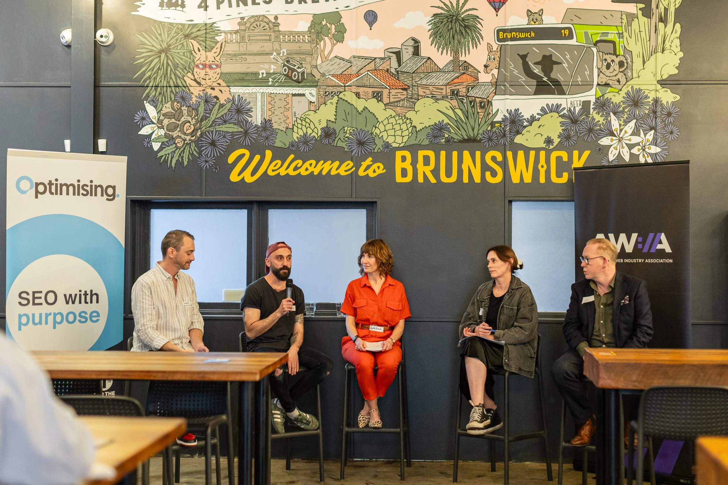 Panel discussion at an event with five people sitting on a stage in front of a large mural that reads 'Welcome to Brunswick'. The mural depicts plants, flowers, and cartoon animals, including a fox and an owl, along with buildings and a bus. Two vertical banners are visible, one for 'Optimising' and the other for the Web Industry Association.