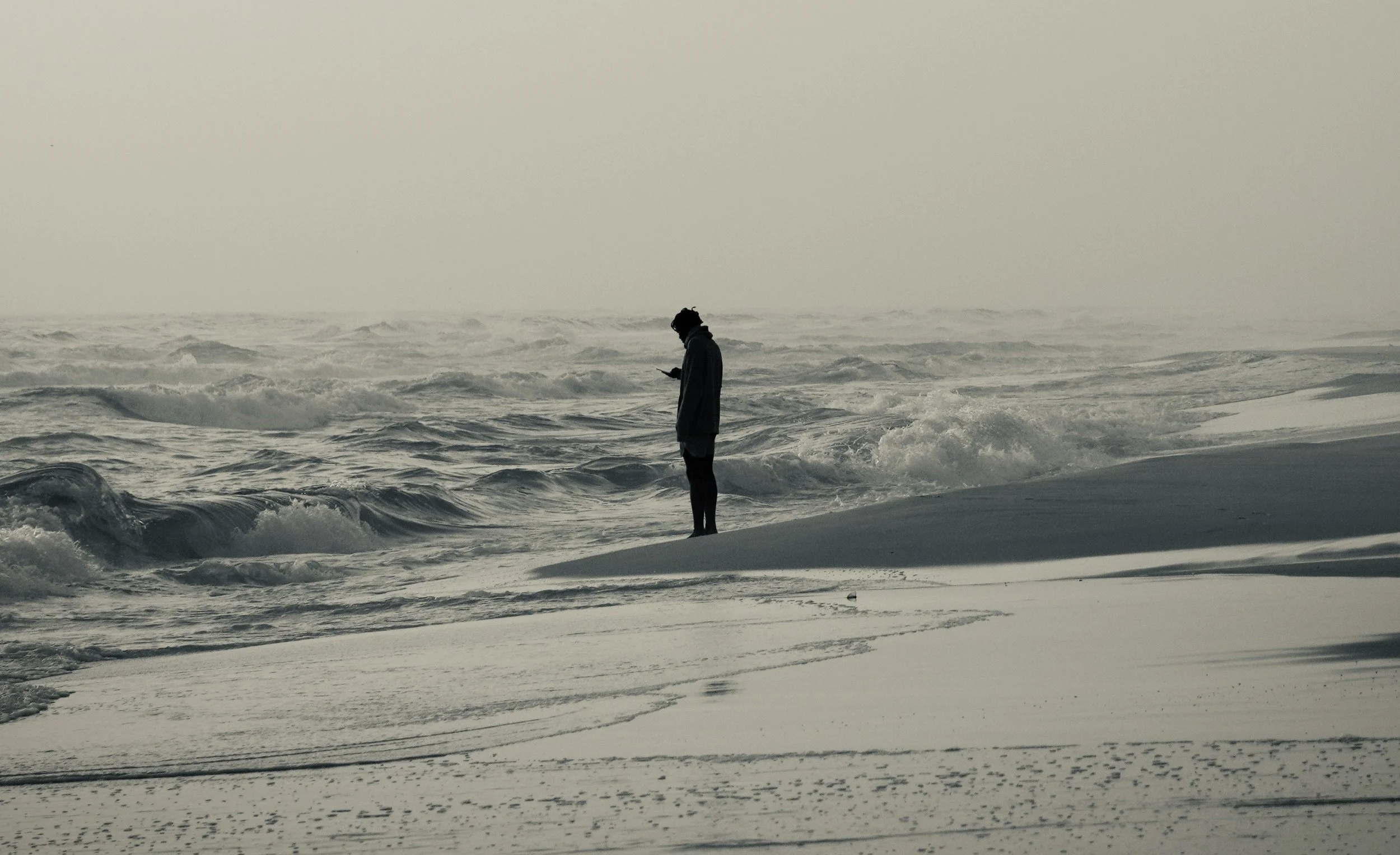 Person in dark clothing standing on a snowy beach, looking at their phone with large ocean waves in the background.