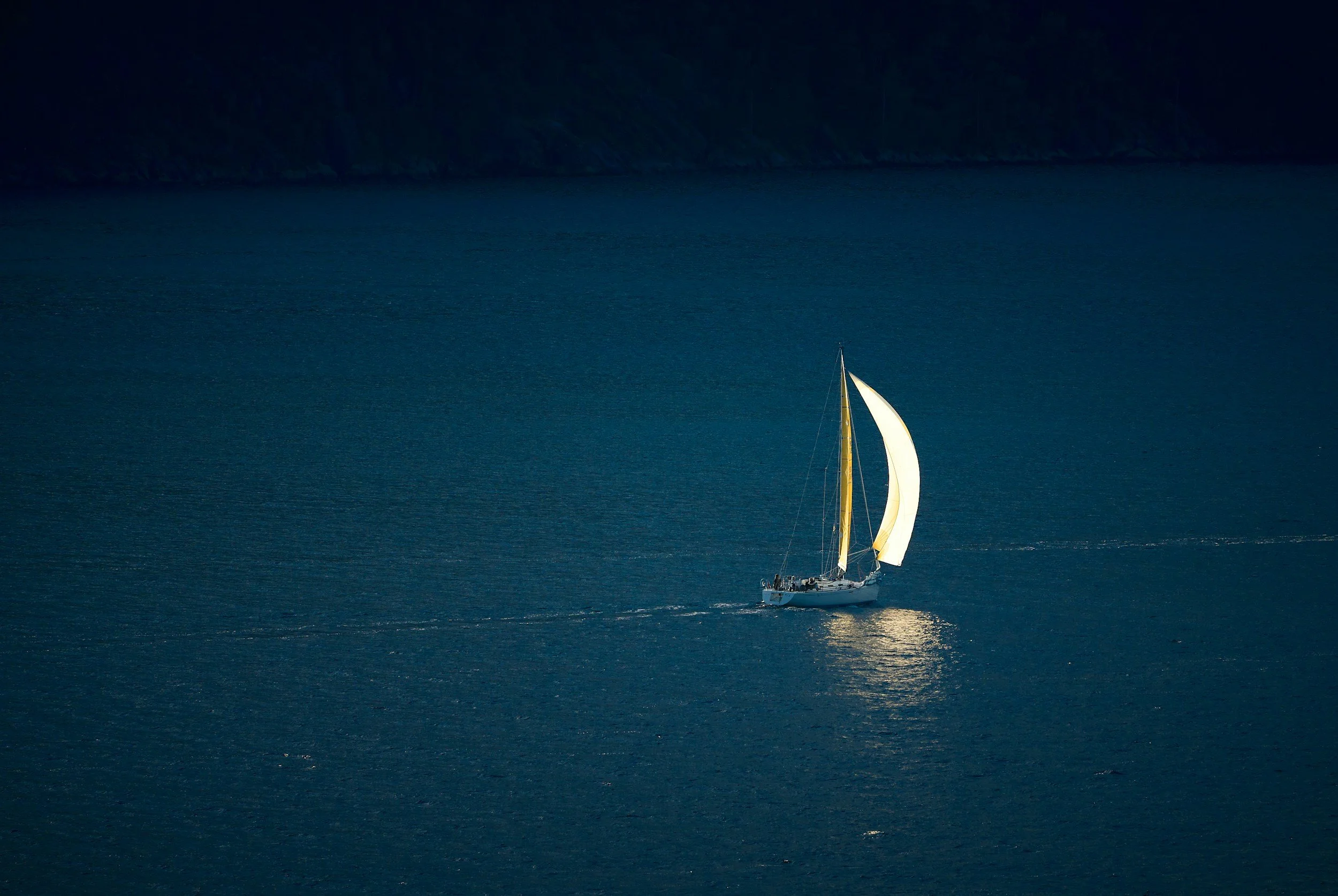 A sailboat with white and yellow sails on calm blue water, with a dark landmass in the background.