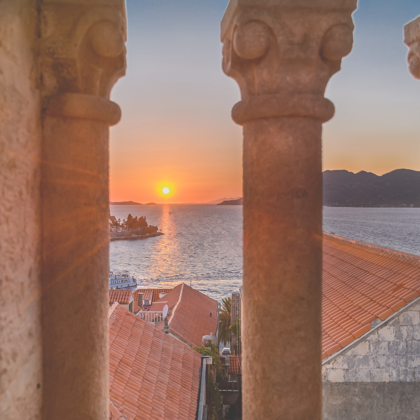 Sunset over the ocean in Korčula viewed through stone pillars and traditional rooftops