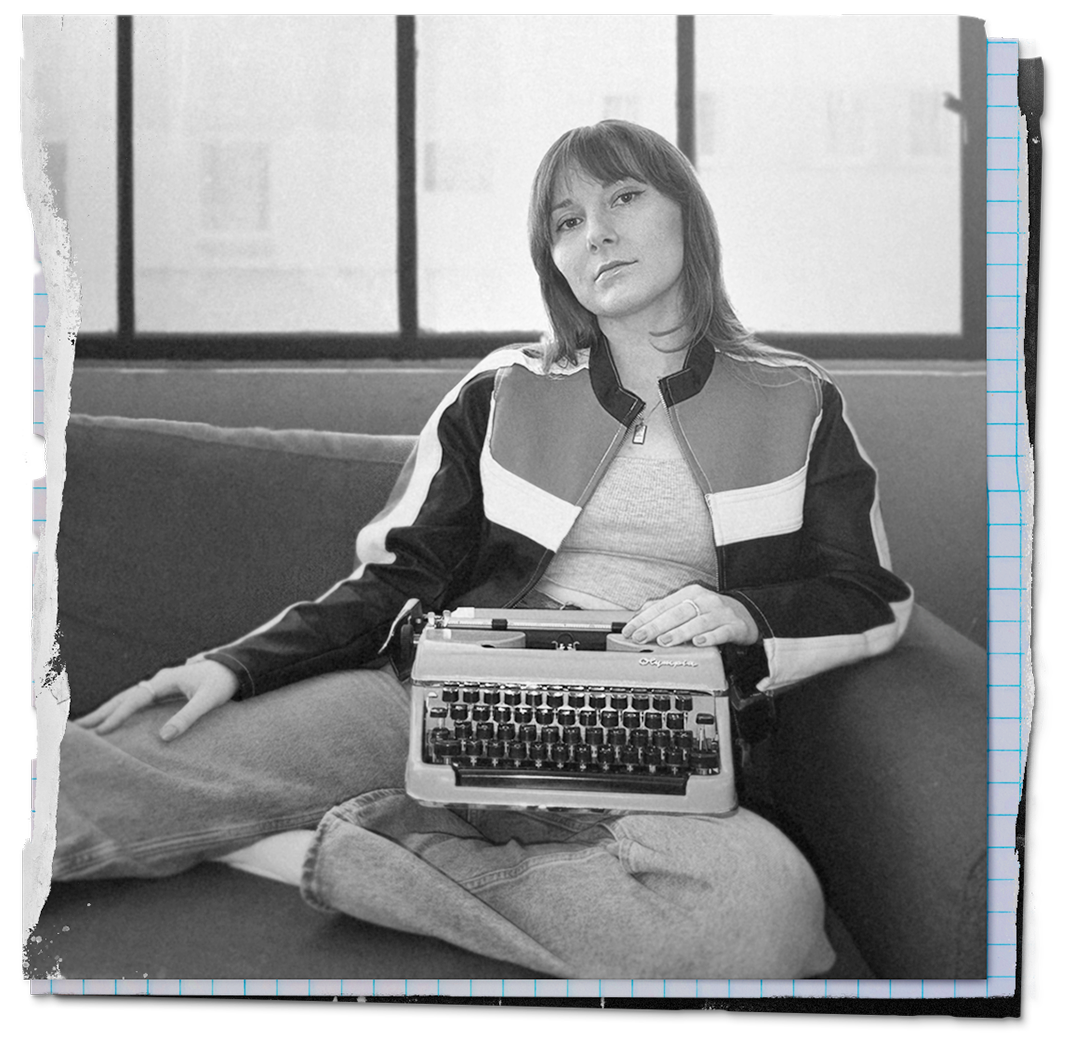 A woman with shoulder-length hair sitting on a couch holding a vintage typewriter on her lap, in front of a large window. The photo is in black and white.