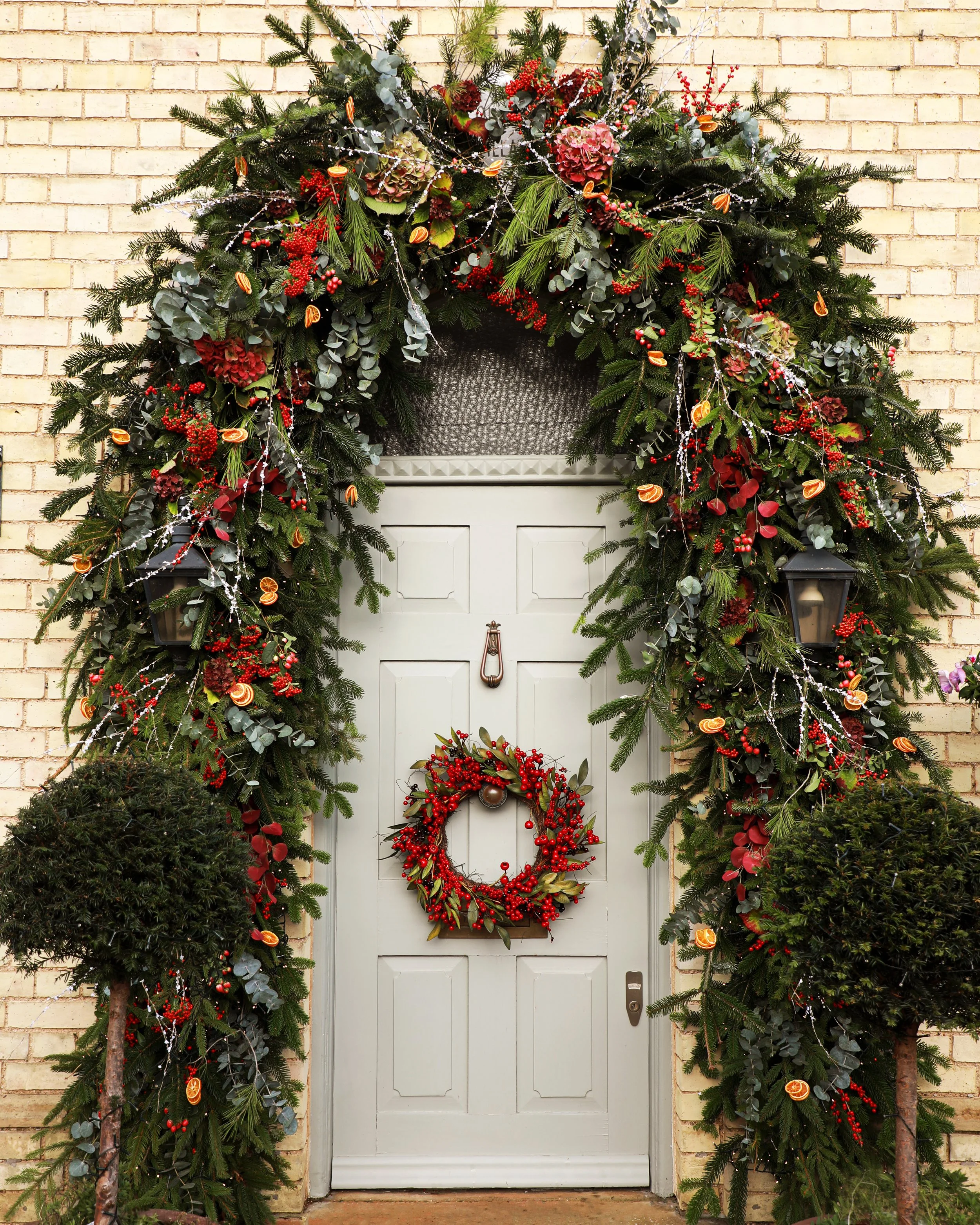 Decorated front door with a Christmas wreath hung on the door and a festive garland arch around it, featuring greenery, red berries, orange slices, and lights, with two potted trees on either side and brick walls in the background.