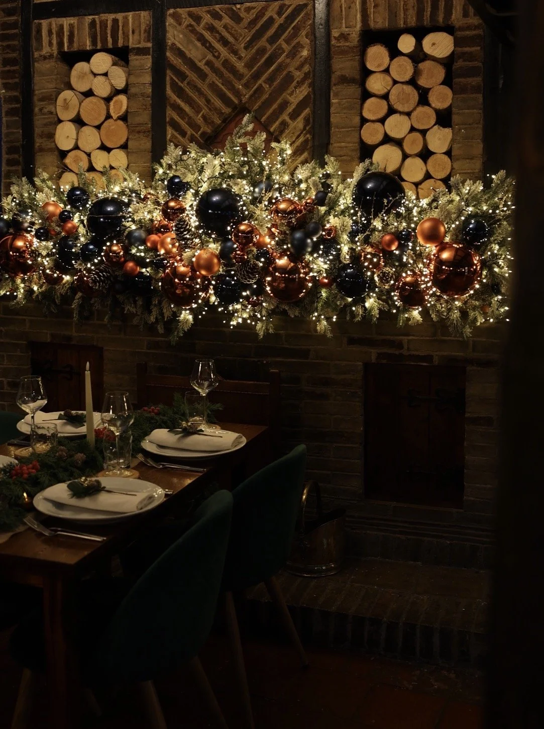 A decorated fireplace mantel with Christmas ornaments, pinecones, and lights, above a dining table set for a holiday meal in a cozy, warmly lit room.
