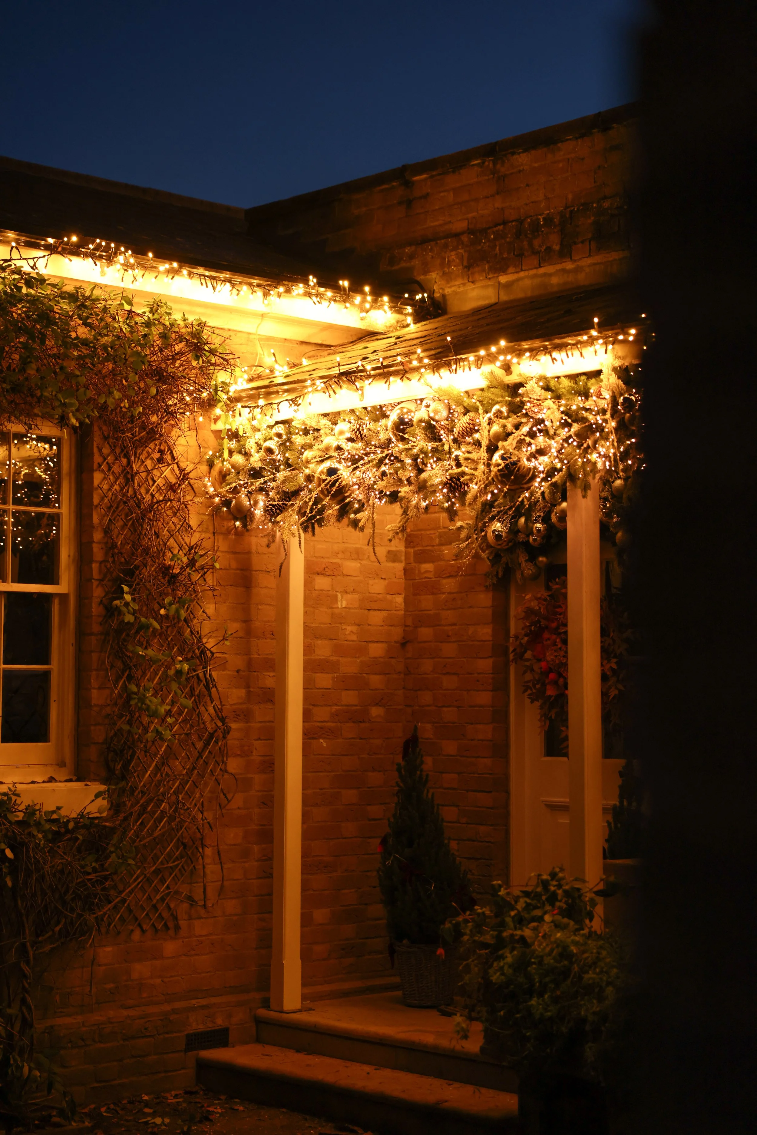 Decorated porch entrance with Christmas lights, ornaments, and greenery at night.