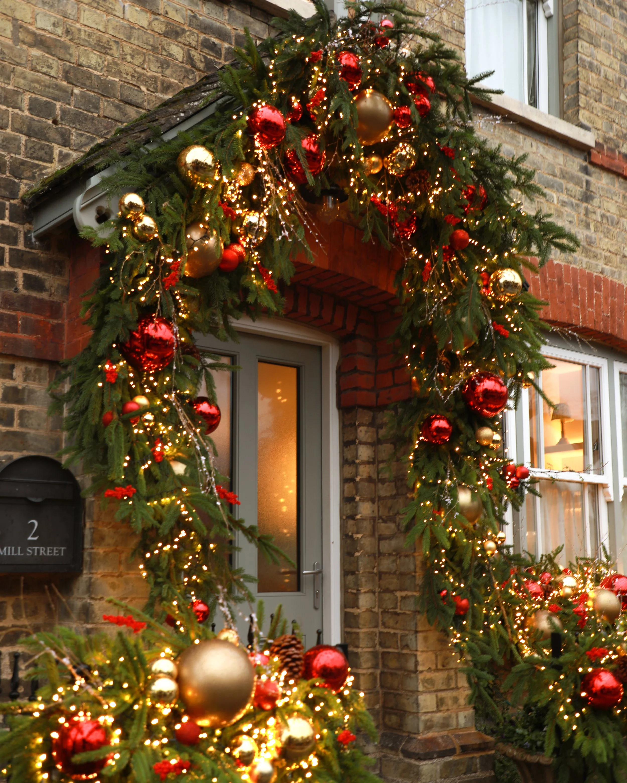 Rustic Christmas decorations with green pine branches, red and gold baubles, and warm lights framing the front door of a period house.