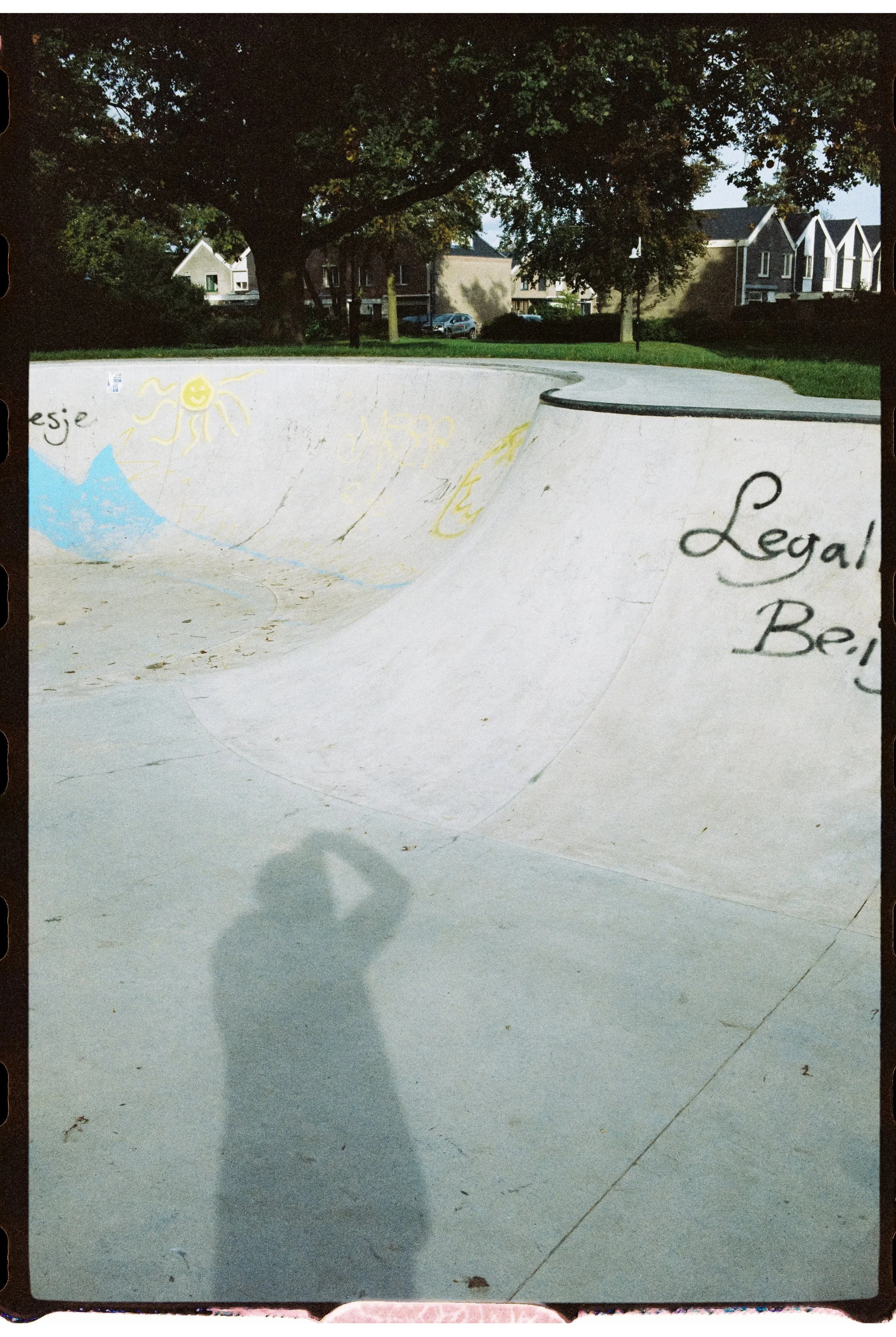 Ein leeren Skatepark mit Graffiti an der Wand, umgeben von Bäumen und Häusern im Hintergrund, mit einem Schatten einer Person, die ein Foto macht.