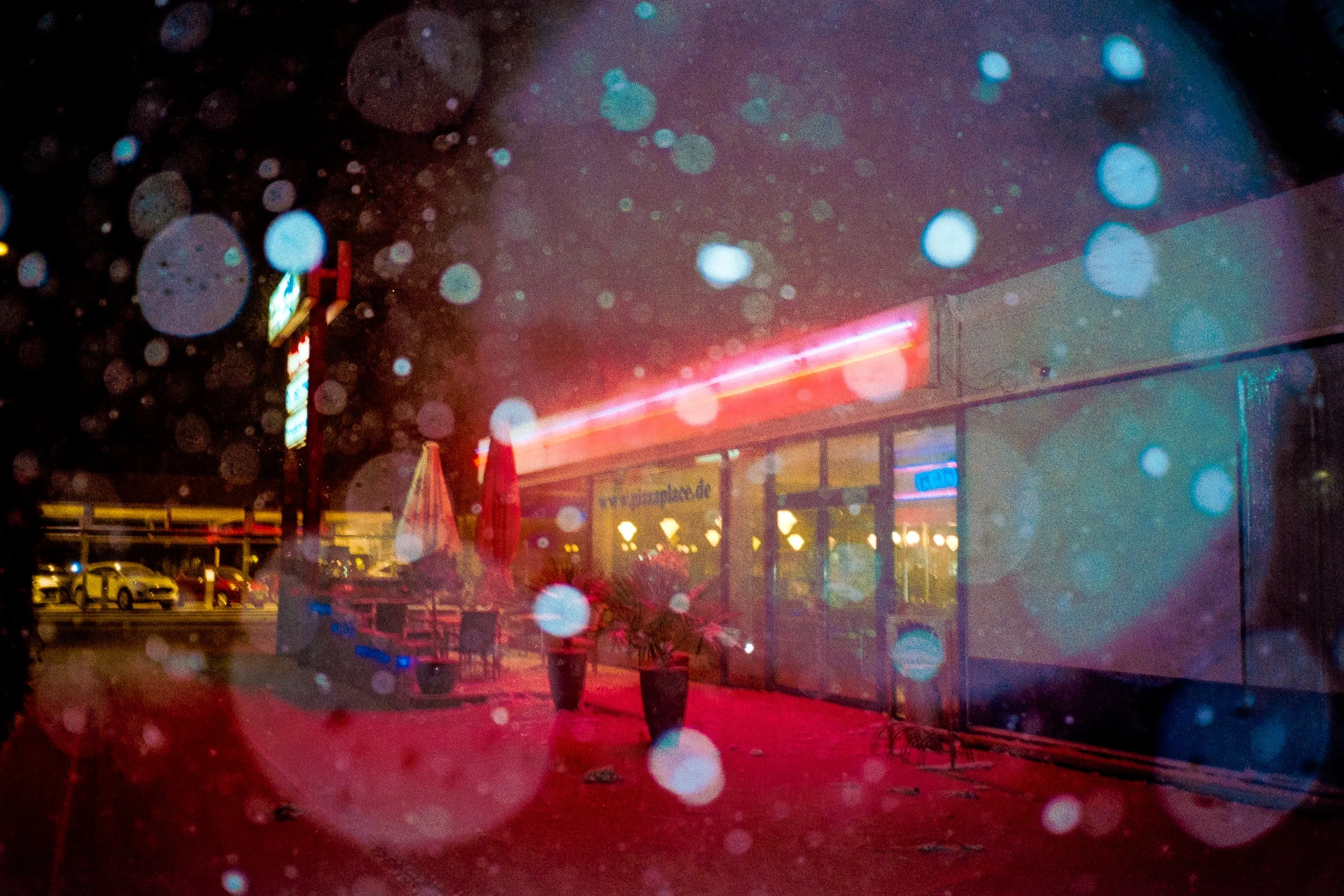 Abendaufnahme einer Außenterrasse eines Restaurants oder Cafés mit Neonlichtern, eingedeckten Tischen und Sonnenschirmen, durch einen Regen bedingt unscharf. Im Hintergrund sind parkende Autos sichtbar.