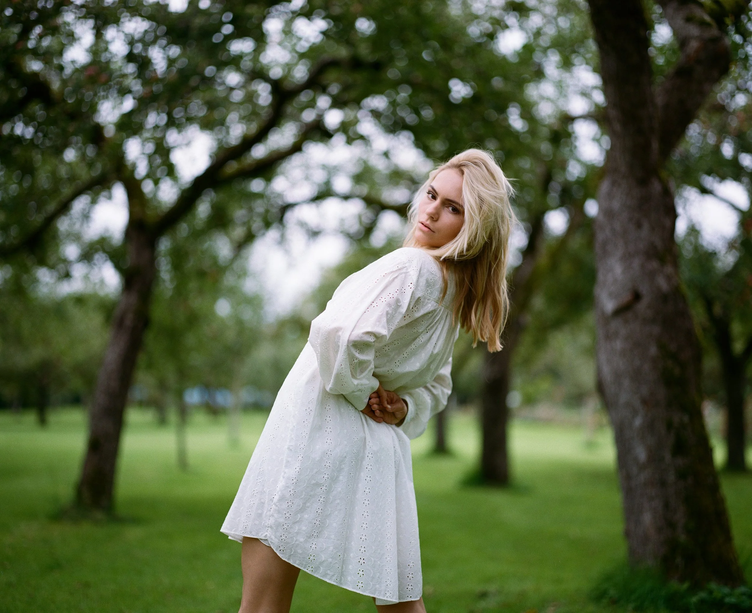 Junge Frau in weißem Kleid in einem Park mit Bäumen im Hintergrund.