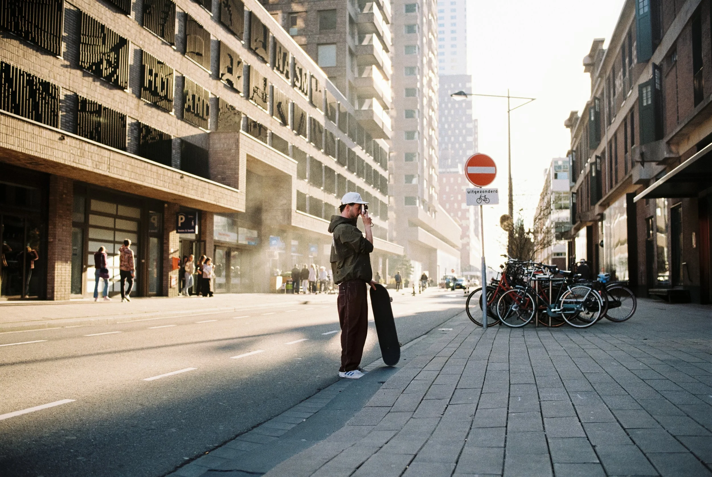 Ein junger Mann mit Skateboard steht an der Straßenseite, spricht im Handyklingel. Im Hintergrund stehen Leute auf dem Gehweg, Bäume, Fahrradständer mit Fahrrädern, und Hochhäuser in einer Stadtumgebung bei Tageslicht.