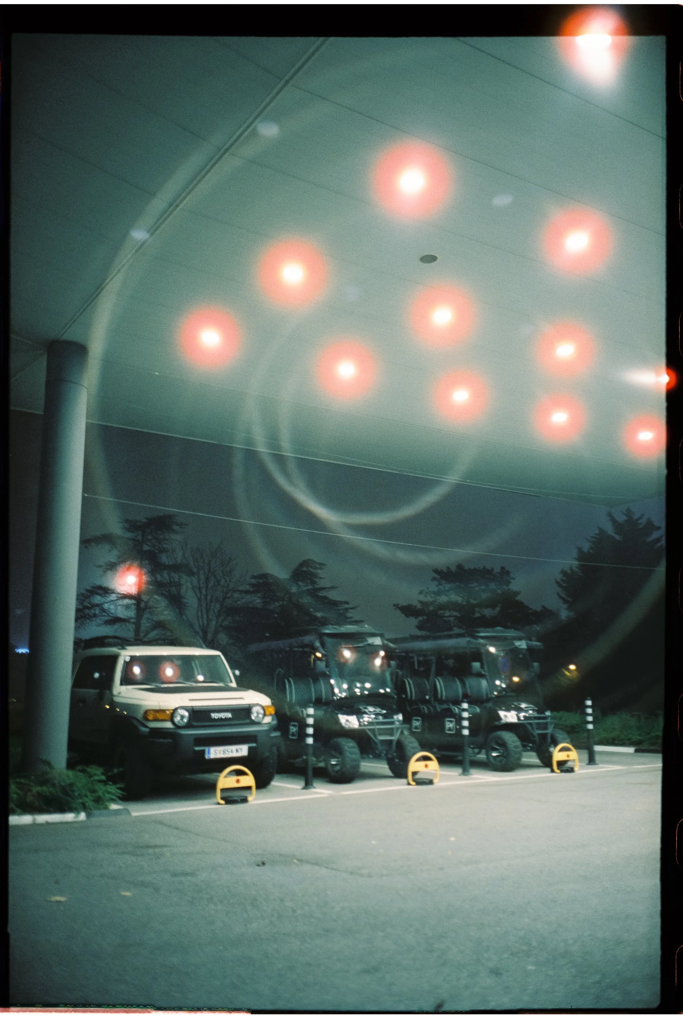 Reflexion der Deckenbeleuchtung auf der Fensterscheibe eines Autos im Parkhaus bei Nacht, mit mehreren geparkten Fahrzeugen und Bäumen im Hintergrund.
