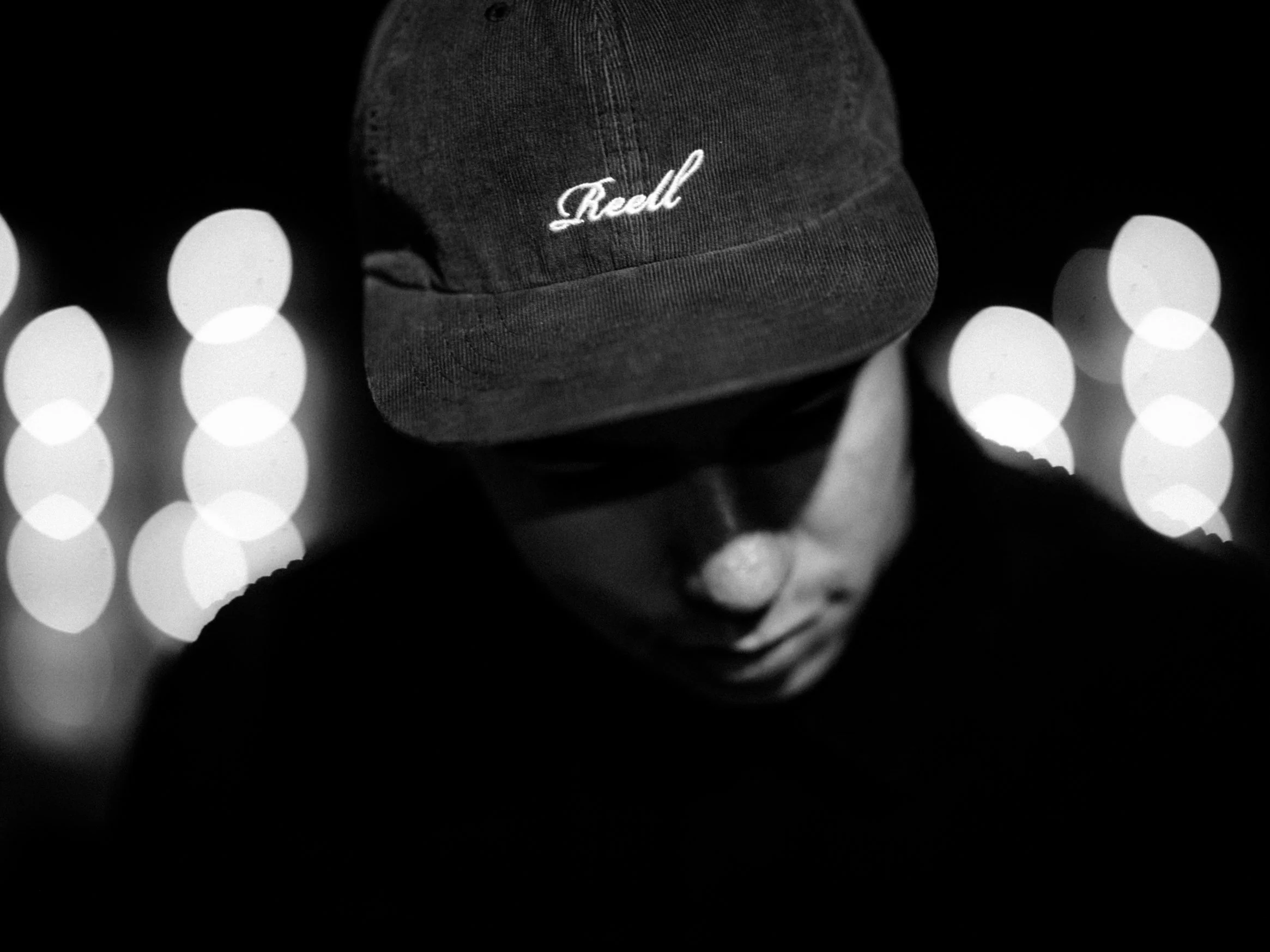Porträt eines Mannes in Schwarz-Weiß, der eine Mütze trägt mit der Aufschrift 'Real', und seinen Kopf leicht nach unten neigt, mit unscharfen Lichtern im Hintergrund.