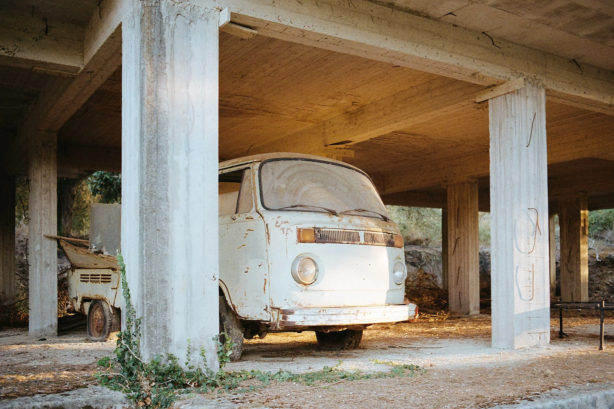 Verrosteter, veralteter Kleinbus unter einer Brücke, umgeben von Pflanzen und Erde.