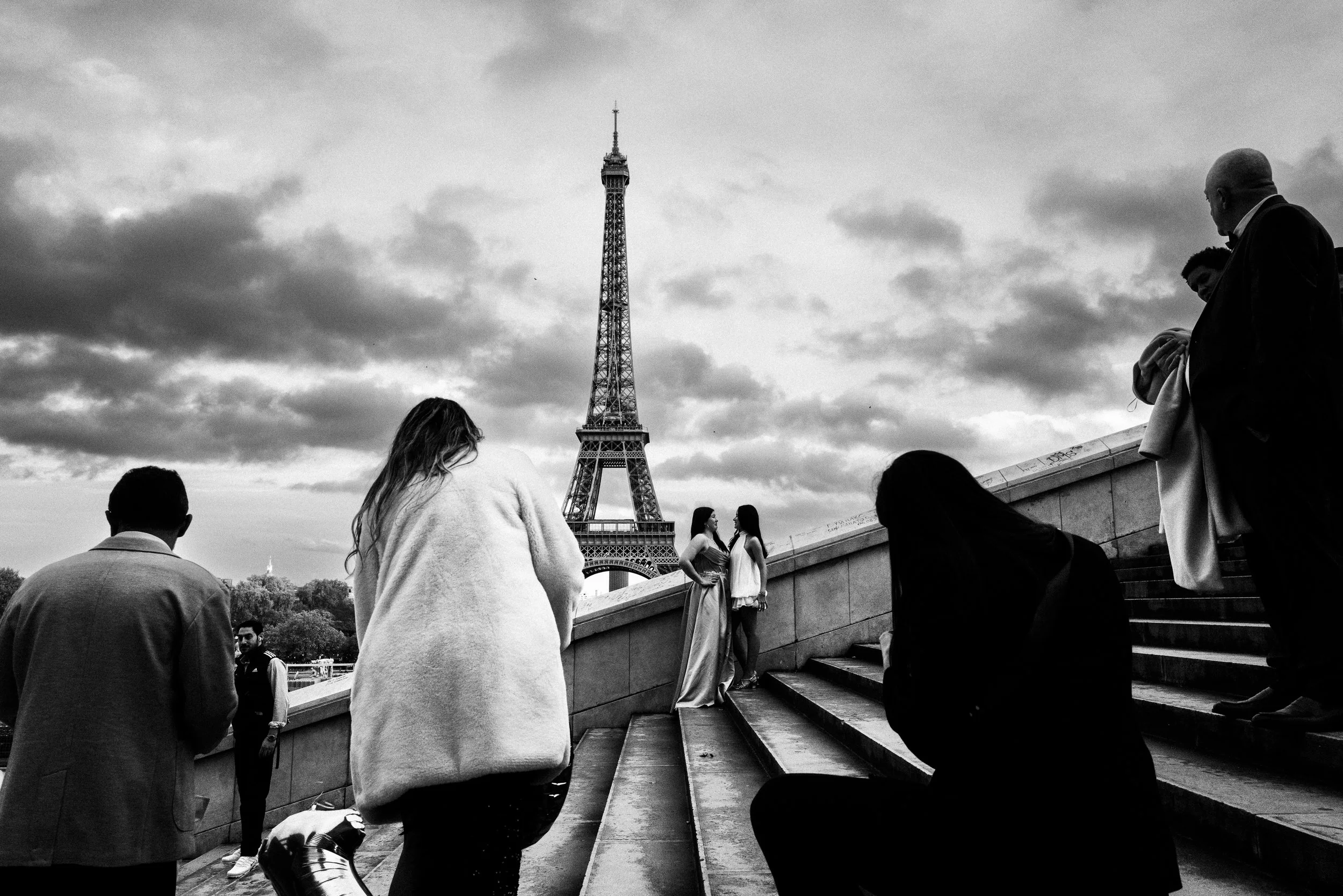 Schwarz-Weiß-Foto von Menschen, die auf den Stufen vor dem Eiffelturm in Paris sitzen und posieren, einige stehen im Hintergrund und schauen.