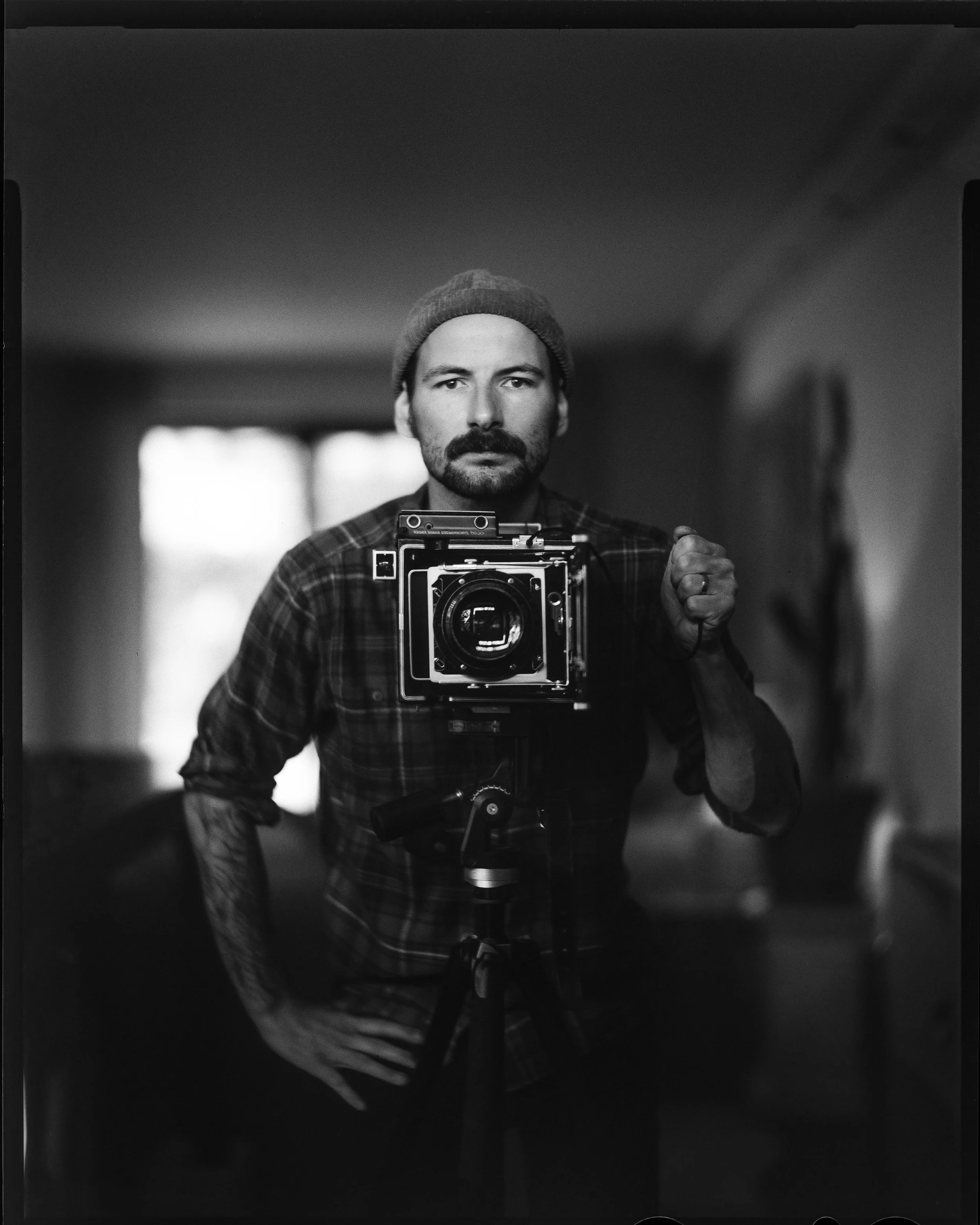 Schwarz-weiß-Foto eines Mannes, der eine Kamera auf einem Stativ trägt, vor einem Spiegel. Er trägt ein Wollmütze und ein kariertes Hemd, steht in einem Raum mit unscharfem Hintergrund.