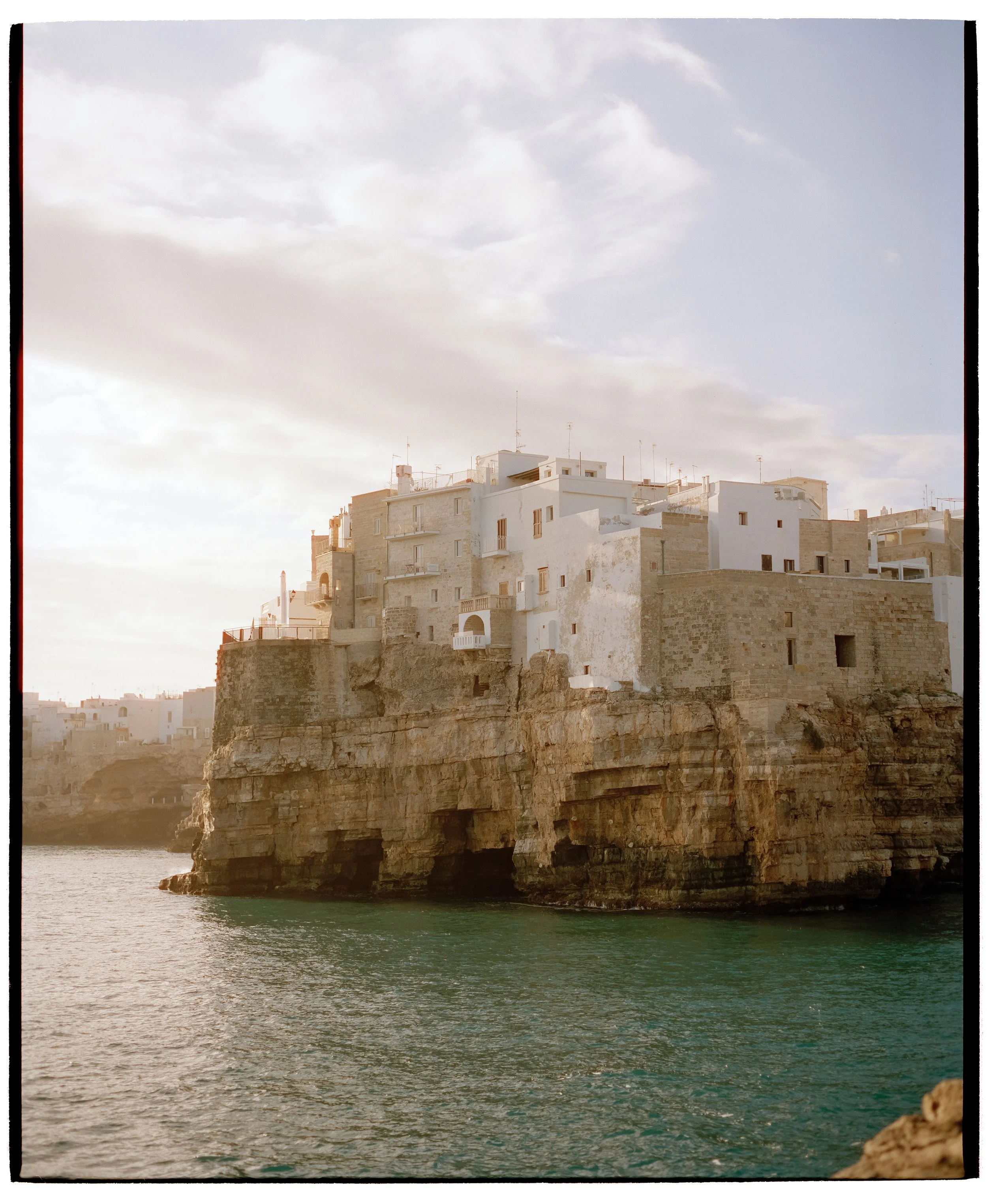 Weiße Häuser auf Felsen vor dem Meer, im Sonnenlicht, mit bewölktem Himmel.