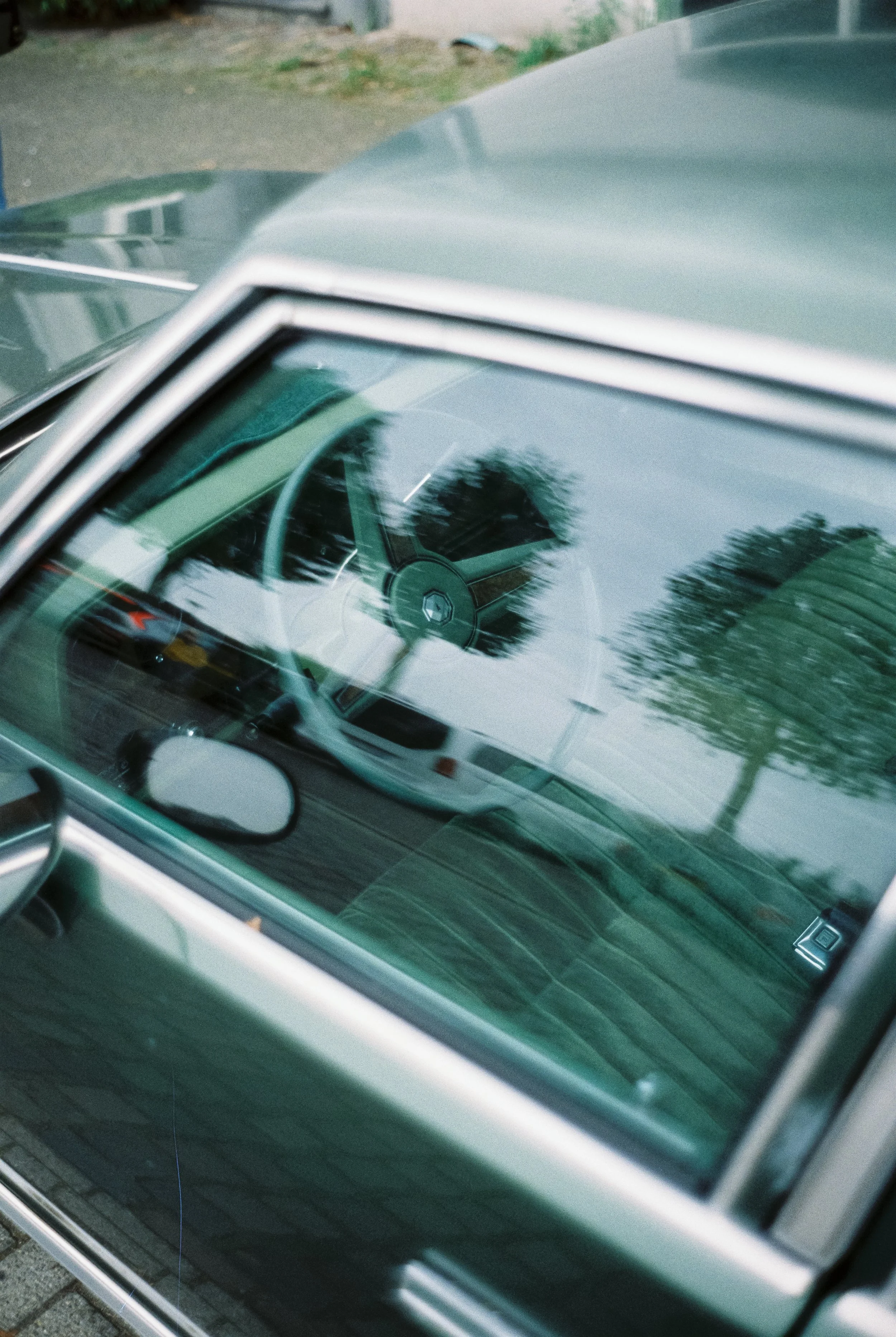 Innenraum eines klassischen Autos mit sichtbarem Lenkrad und Spiegel, im Spiegel reflektiert eine Straße mit Bäumen und einem weißen Auto.