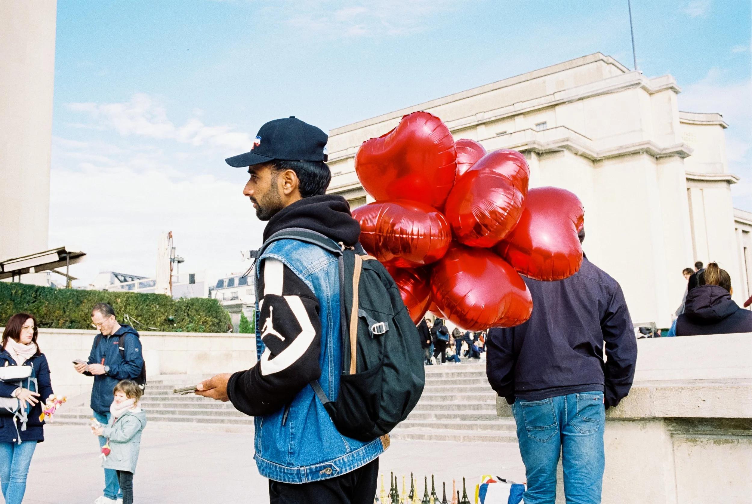 Mann mit schwarzen Ballons in Herzform in einer Menschenmenge vor einem großen weißen Gebäude in Paris.