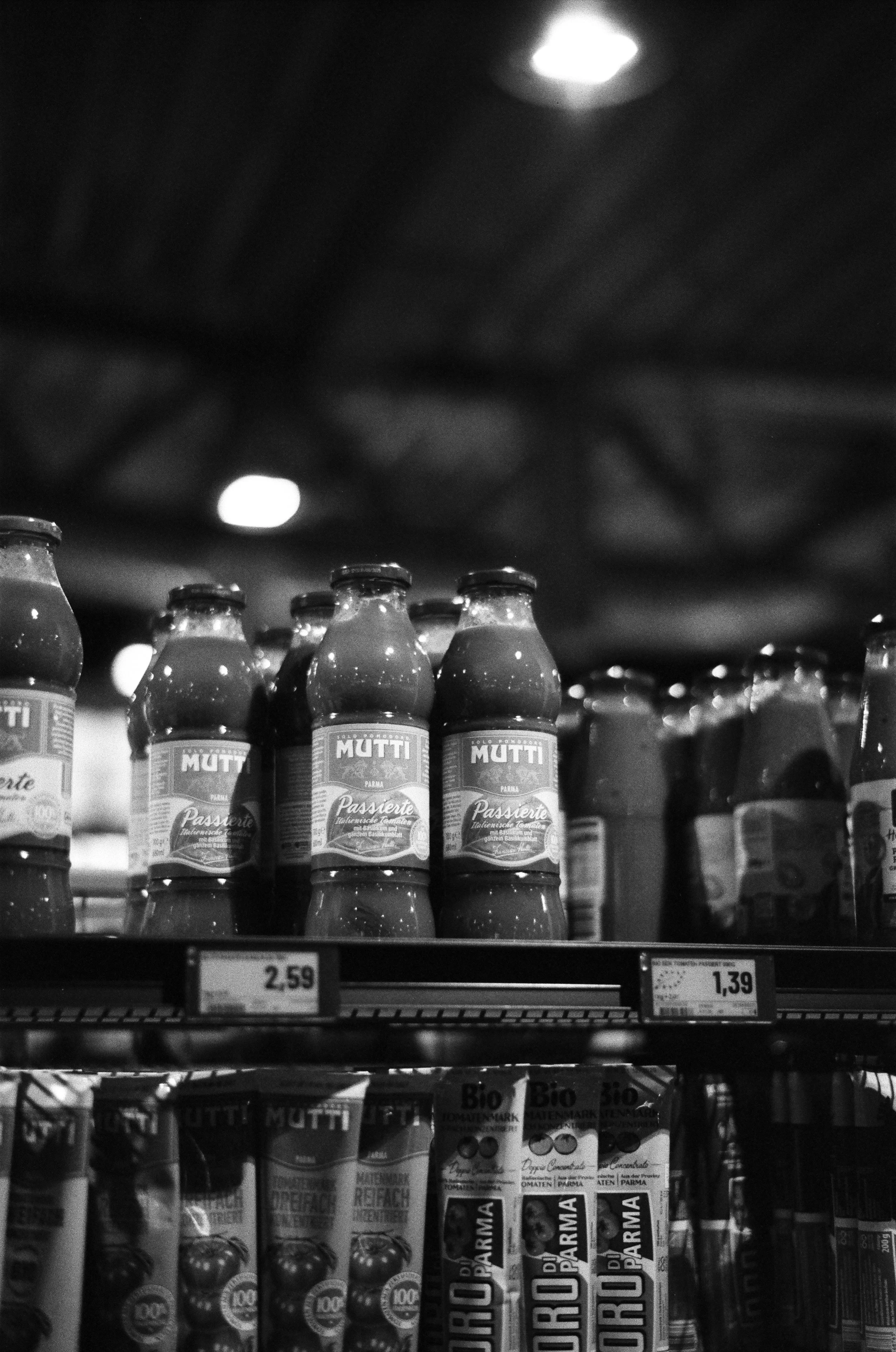 Schwarz-weiß-Foto von gefüllten Flaschen mit Tomatensauce im Supermarktregal. Preisetiketten sind sichtbar.