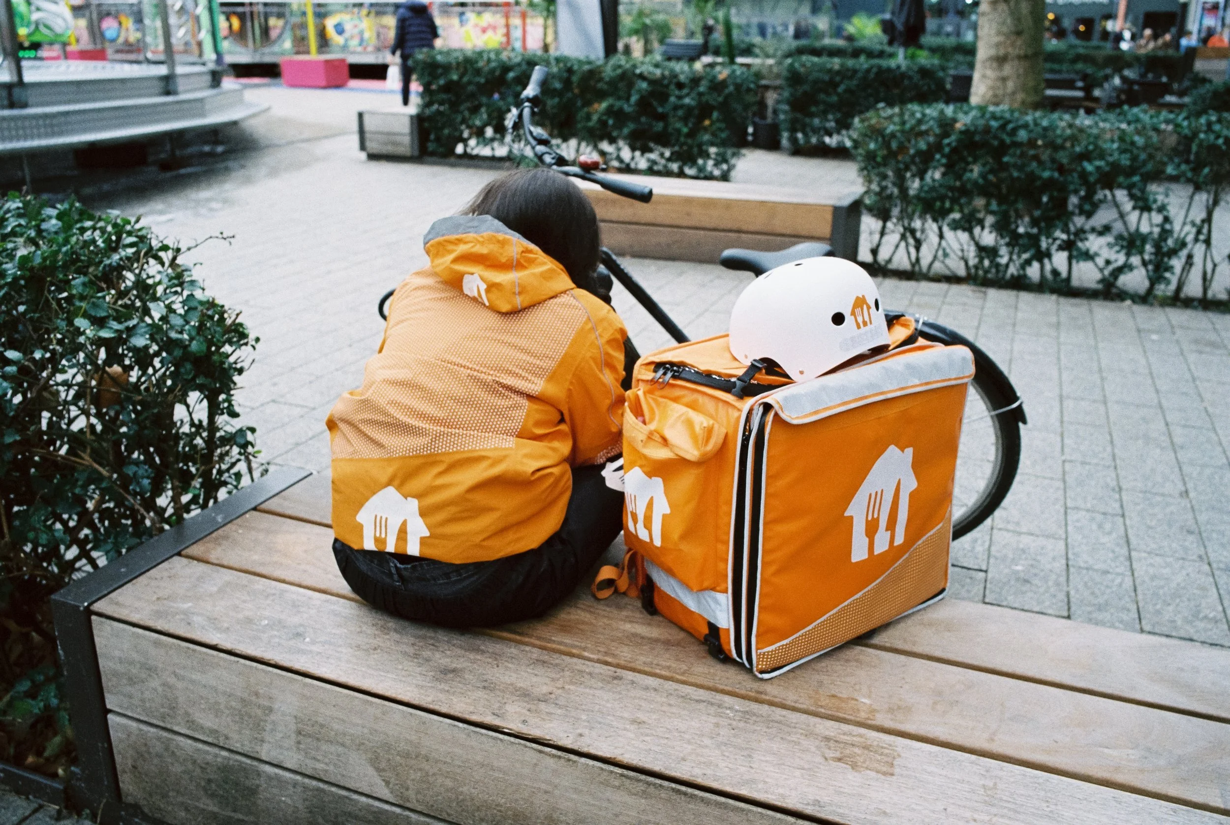 Fahrrad mit gelbem Lieferrucksack und Helm auf einer Parkbank, eine Person in gelbem Jacke sitzt neben dem Fahrrad.