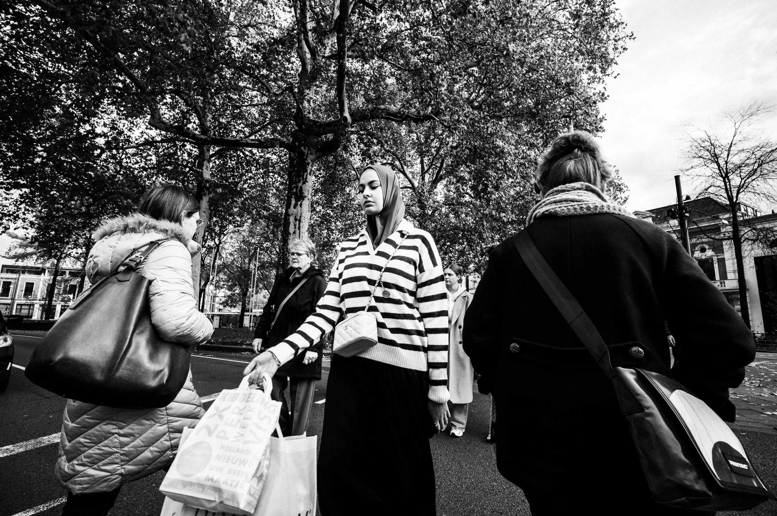 Drei Frauen auf einer Straße, eine trägt einen gestreiften Pullover und einen Hijab, die anderen beiden tragen Winterjacken, im Hintergrund Bäume und Gebäude.