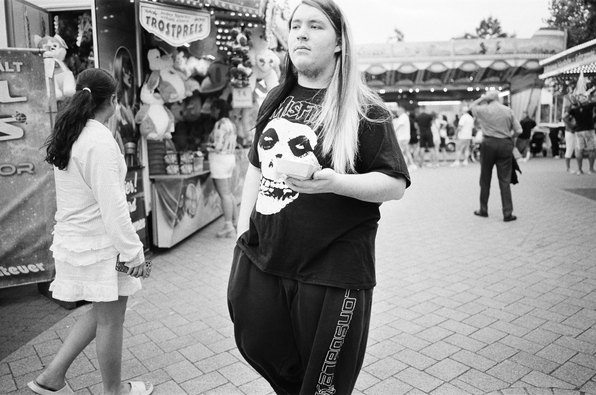 Junge Frau mit langen Haaren in schwarzen T-Shirt mit Totenkopf-Design und schwarzen Hosen steht in einem Jahrmarkt, hält ein Handy in der Hand, andere Leute gehen im Hintergrund.