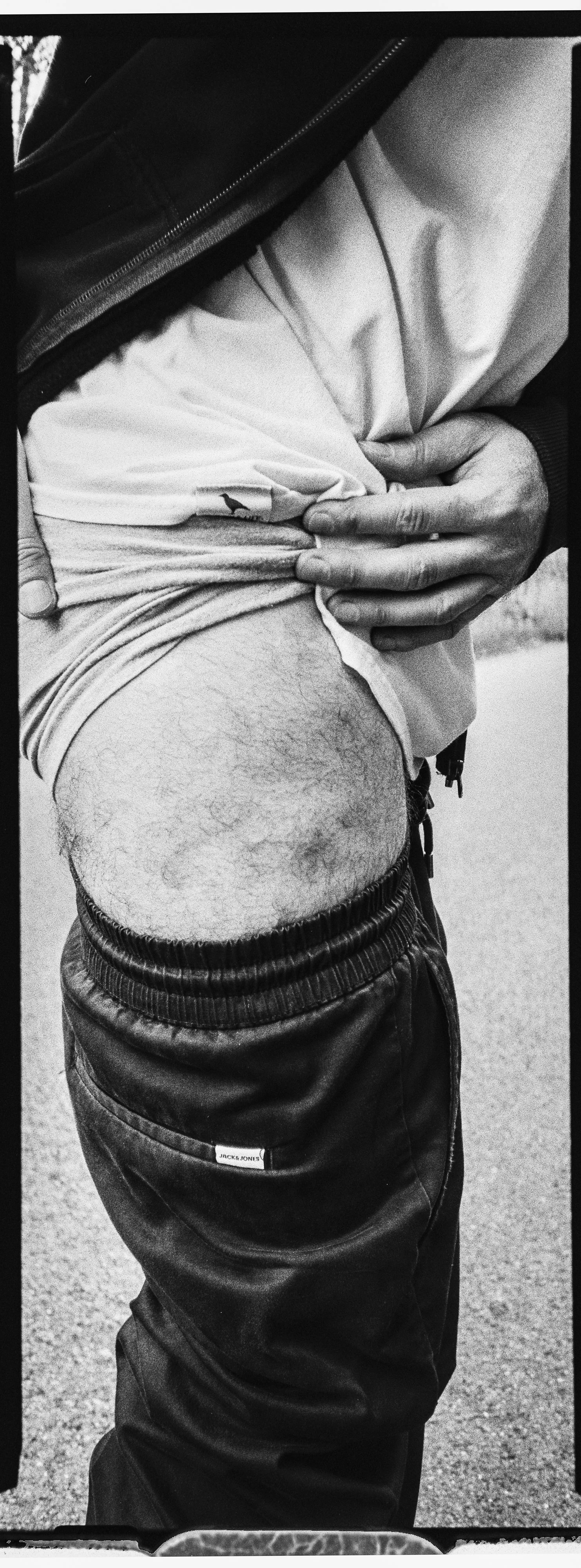 Nahaufnahme eines Mannes, der seine obere Kleidung anhebt, um sein nuda Bauch zu zeigen. Er trägt eine schwarze Jacke, ein weißes T-Shirt und schwarze Jogginghose.