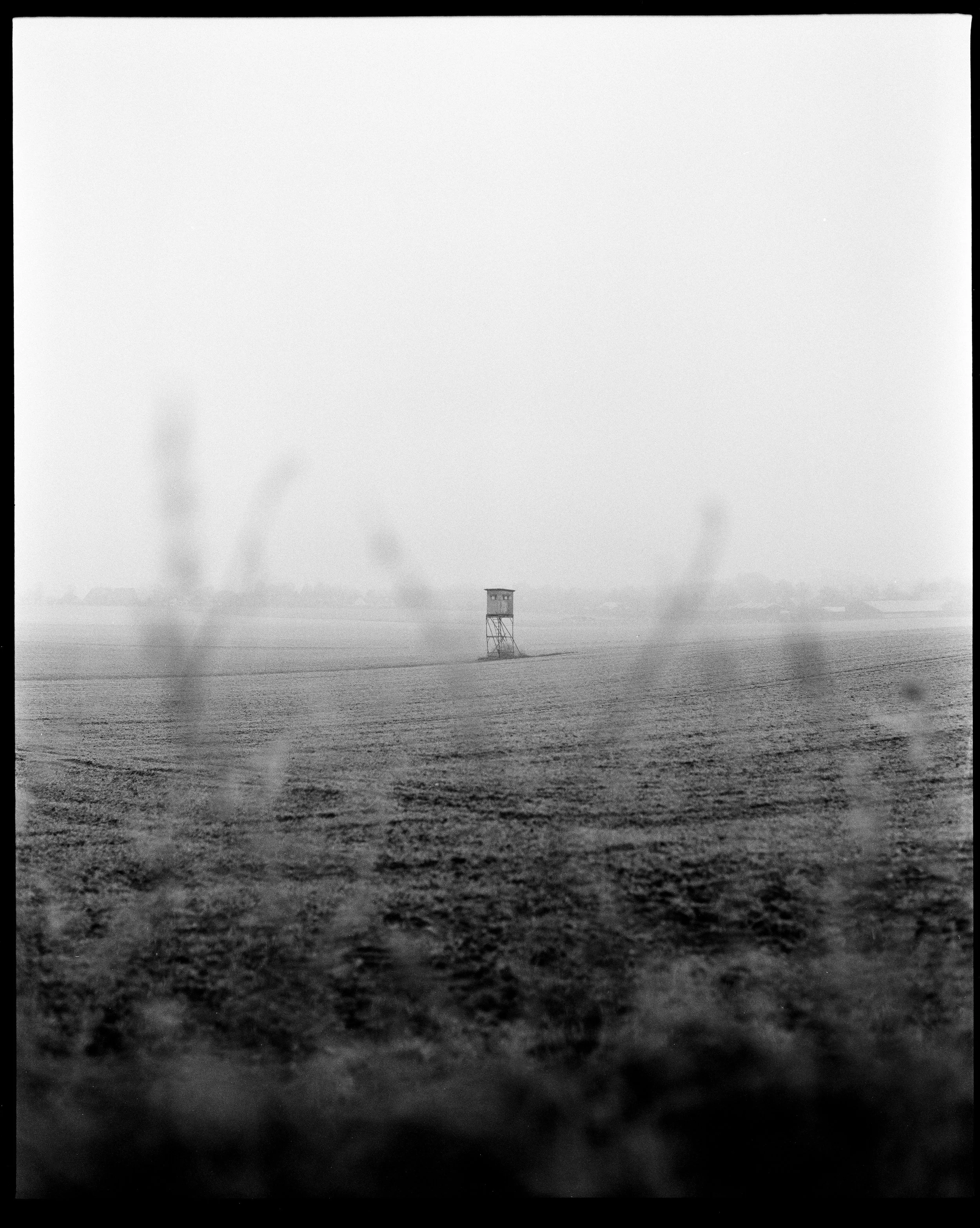 Ein weites, offenes Feld mit einem kleinen Wachturm in der Mitte, leicht neblig und grau, im Hintergrund einige Bäume und eine flache Landschaft.