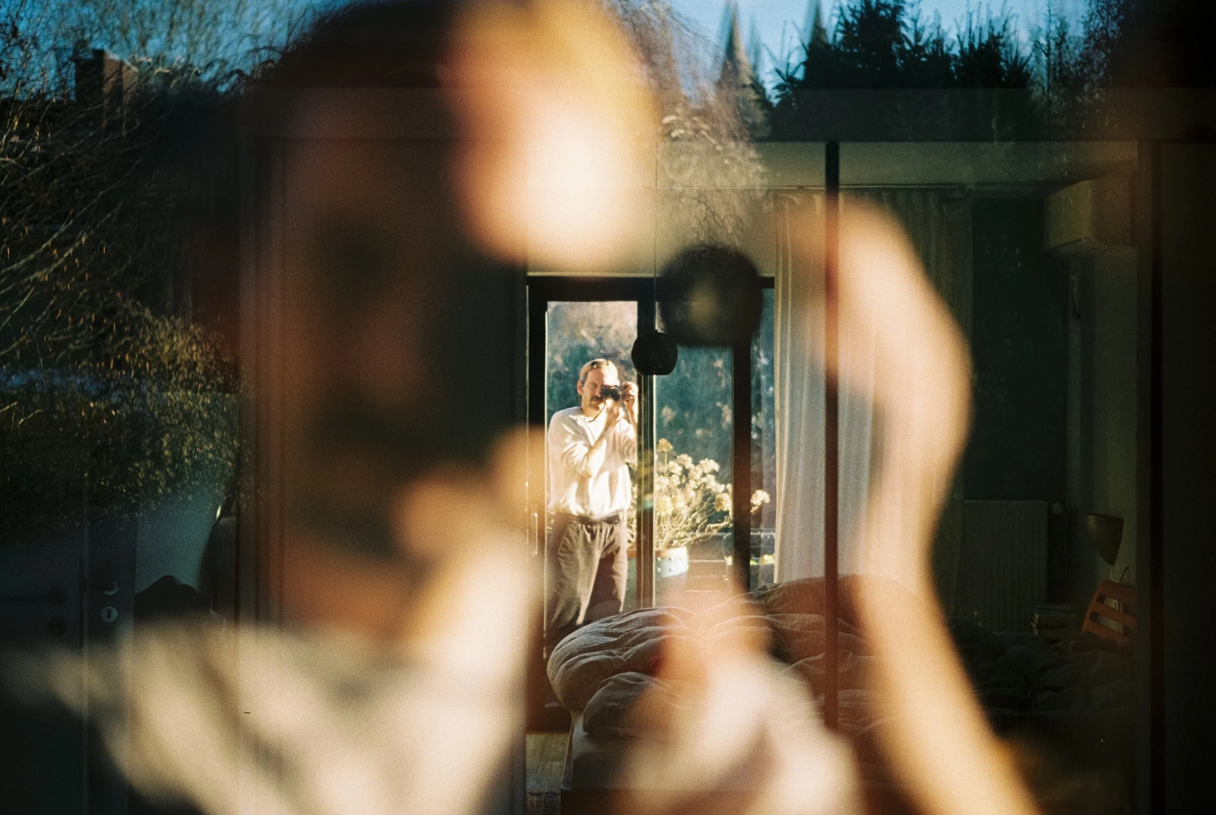 Ein Mann fotografiert sich selbst im Spiegel, während die Kamera durch eine Tür oder Fenster sichtbar ist.