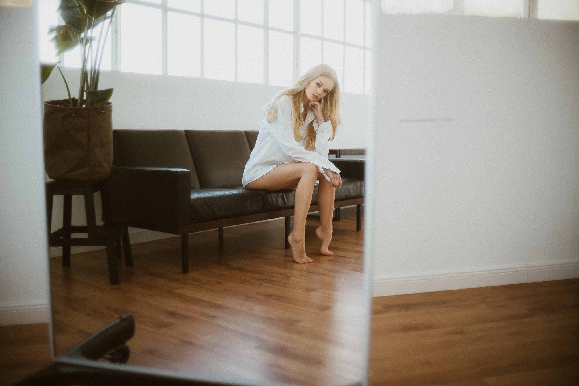 Eine blonde Frau sitzt nachdenklich auf einem schwarzen Ledersofa in einem hellen Raum, während sie in den Spiegel schaut.