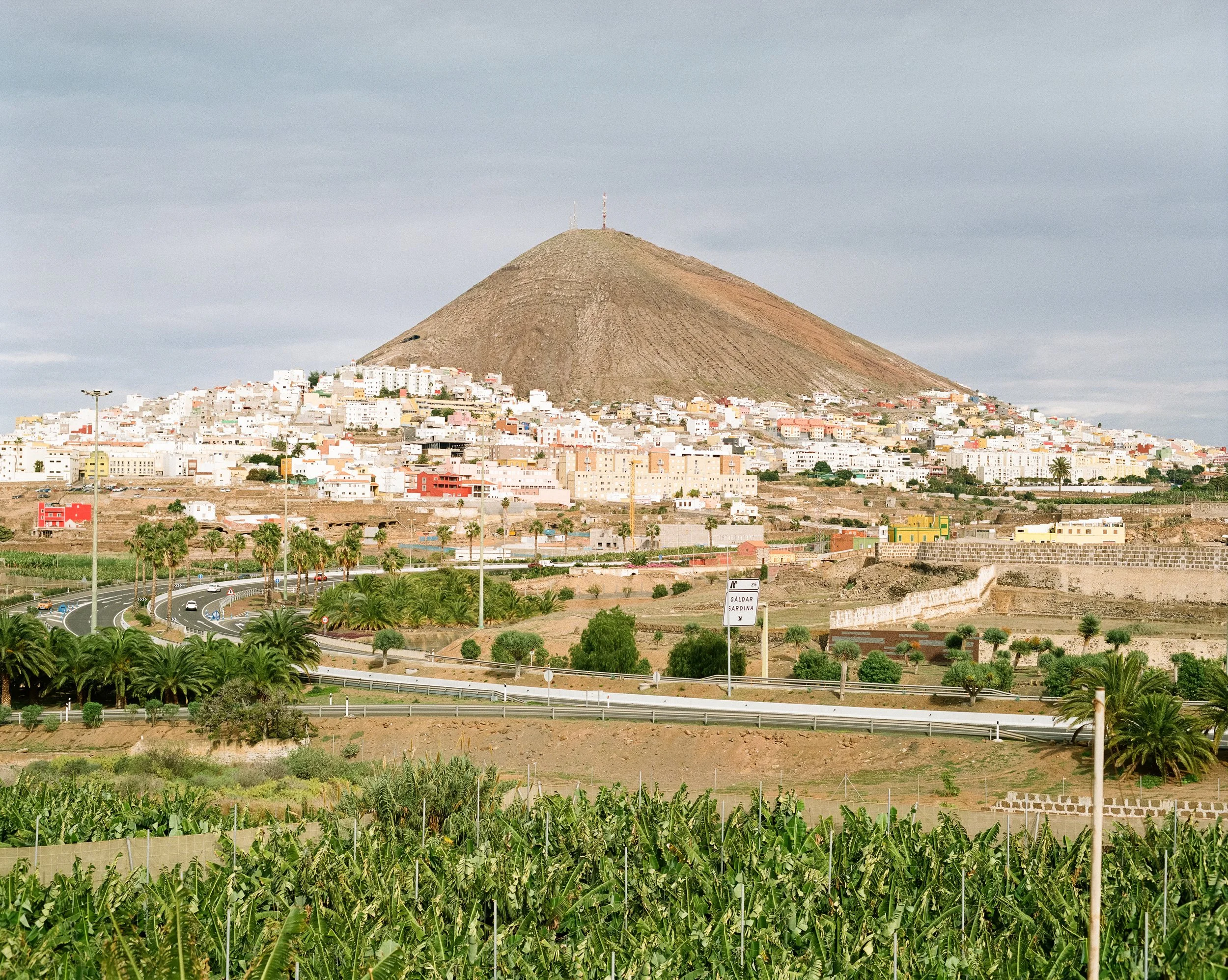 Blick auf die Stadt Galdar in Gran Canaria mit der vulkanischen Silhouette des Vulkans Montaña de Gáldar im Hintergrund, grüne Landwirtschaft im Vordergrund und bewölkter Himmel.
