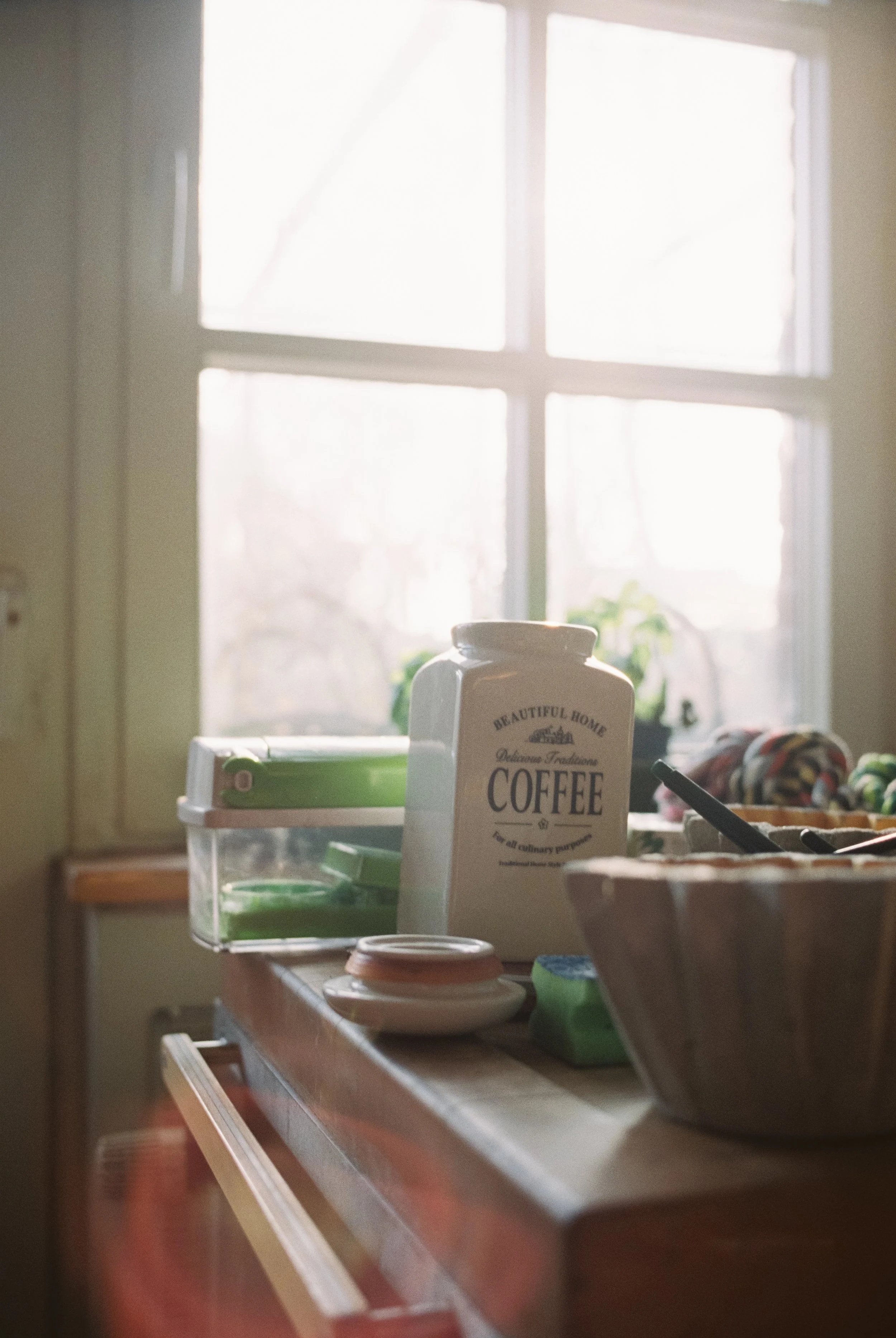 Küchenarbeitsutensilien auf einem Holztisch, im Hintergrund ein Fenster mit natürlichem Licht, ein Keramikbehälter mit Aufschrift "Coffee".