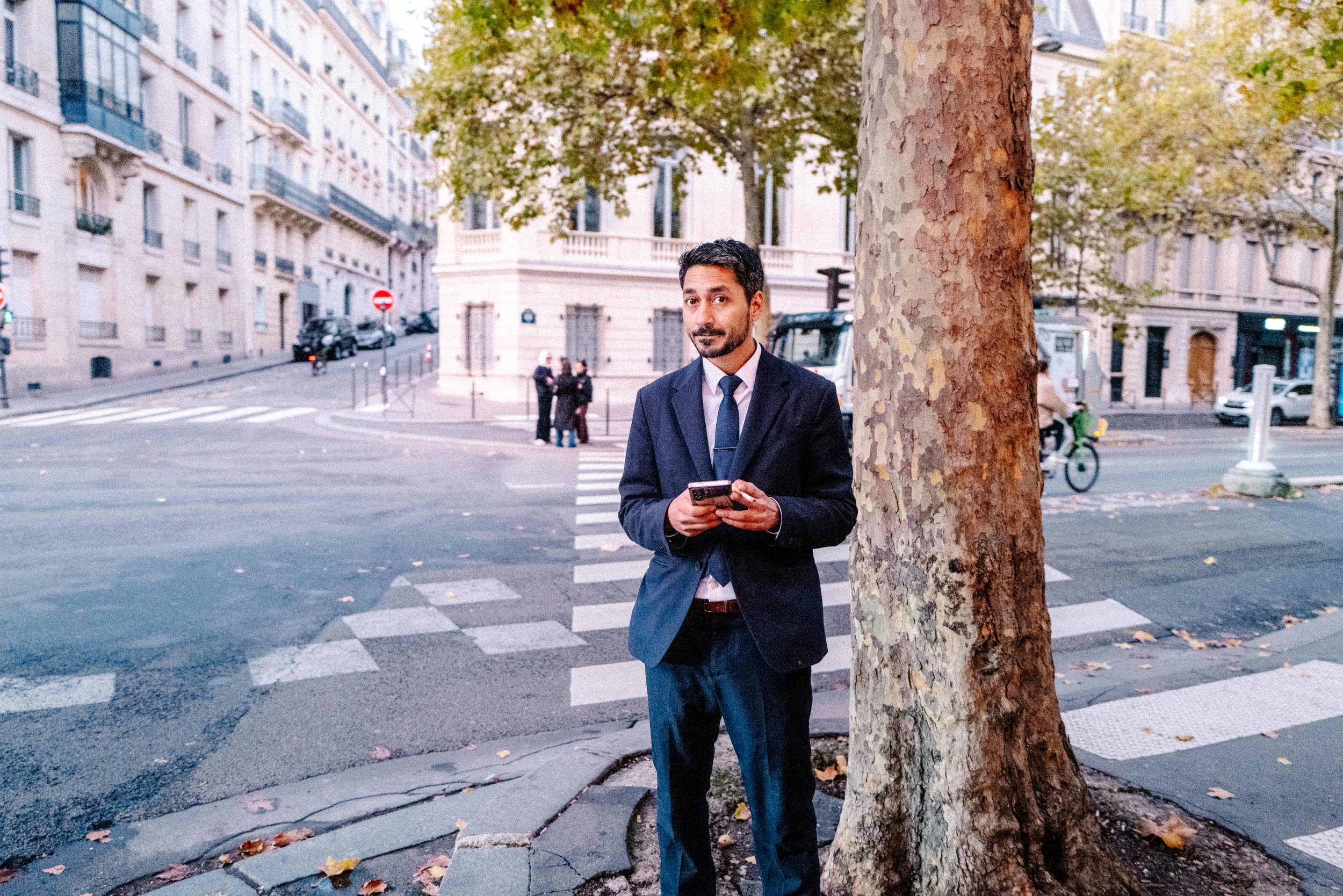 Ein Mann in einem dunklen Anzug steht an einer Straßenkreuzung, hält ein Smartphone, im Hintergrund sind Gebäude, eine Gruppe von Menschen und ein Fahrradfahrer sichtbar.