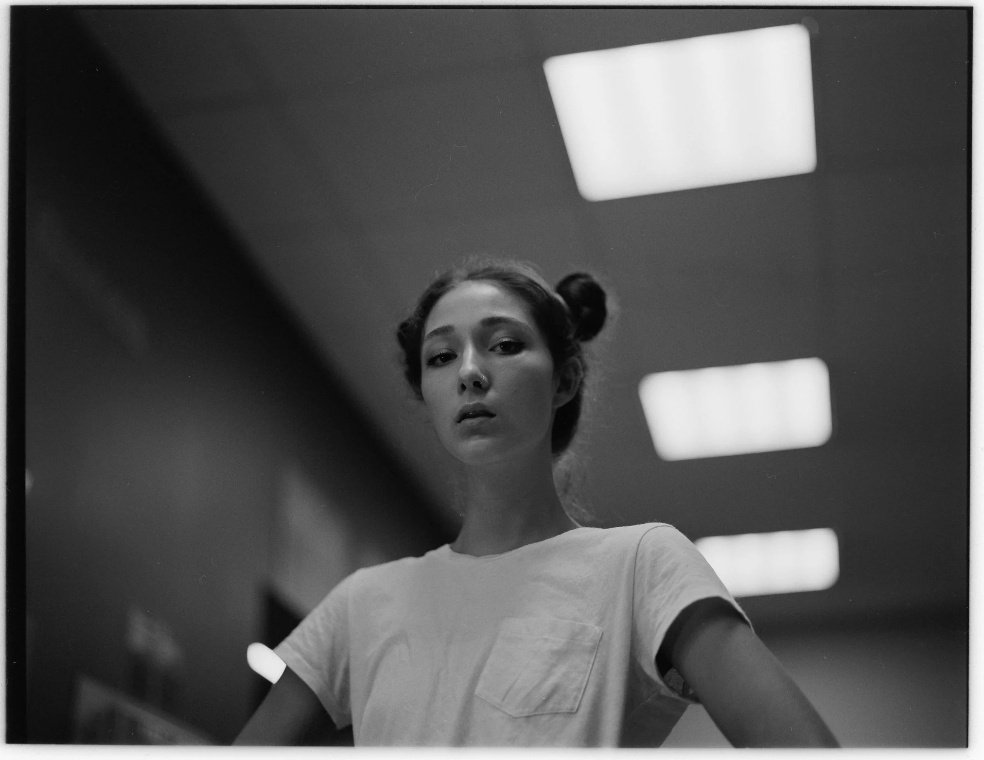 Schwarz-Weiß-Fotografie einer jungen Frau mit zwei Zöpfen, die in den Kamera blickt, im Hintergrund von einem Raum mit Deckenleuchten.