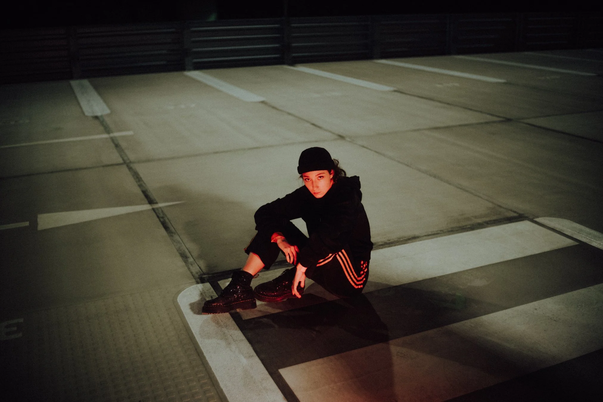 Junge Frau in dunkler Kleidung und schwarzem Hut sitzt in einem leeren, dunklen Parkhaus, beleuchtet von warmem Licht.