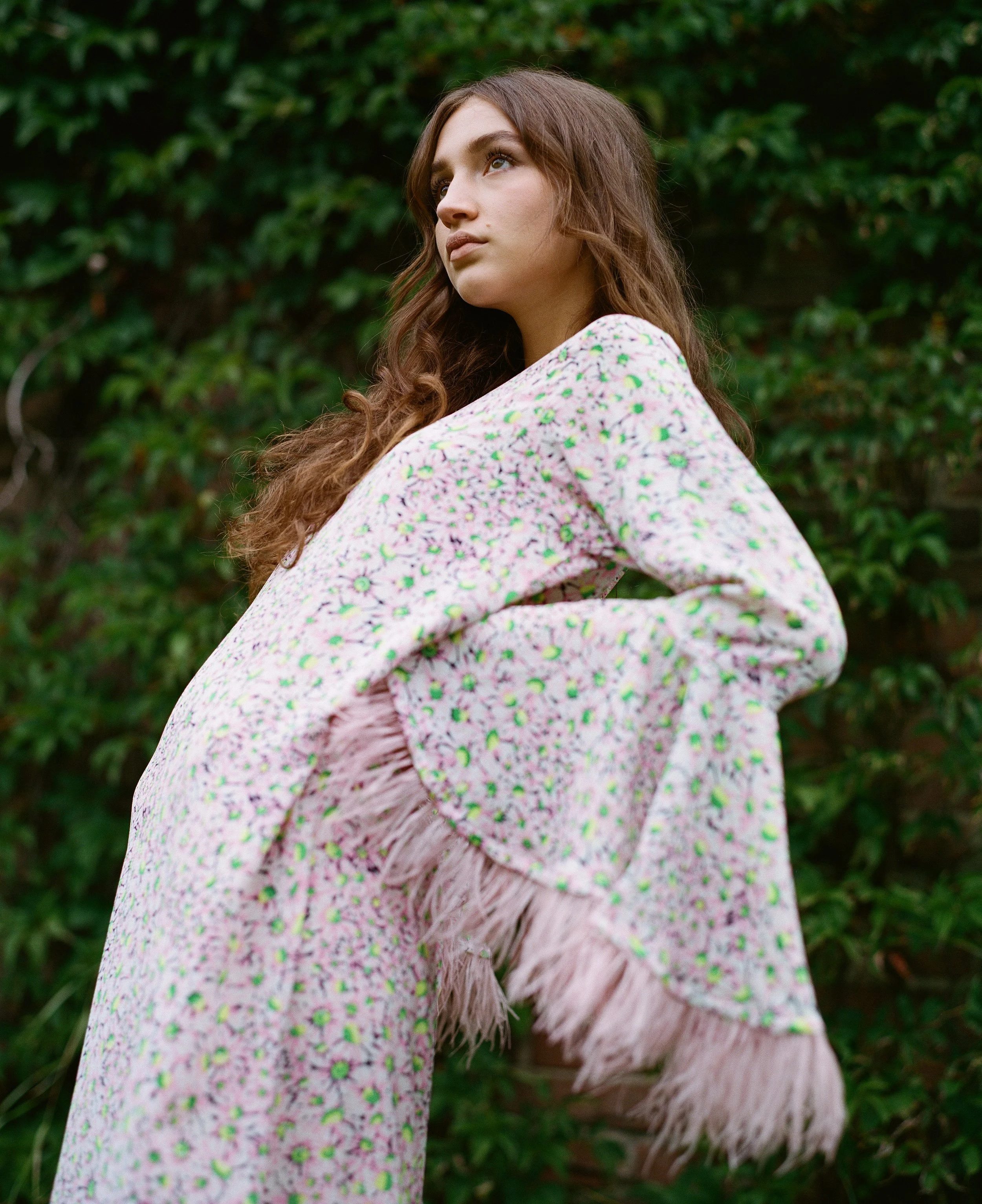 Junge Frau mit langen braunen Haaren trägt ein pinkfarbenes Kleid mit grünen, weißen und lila Blumenmustern und Fransen an den Ärmeln, steht vor einer grünen Hecke.