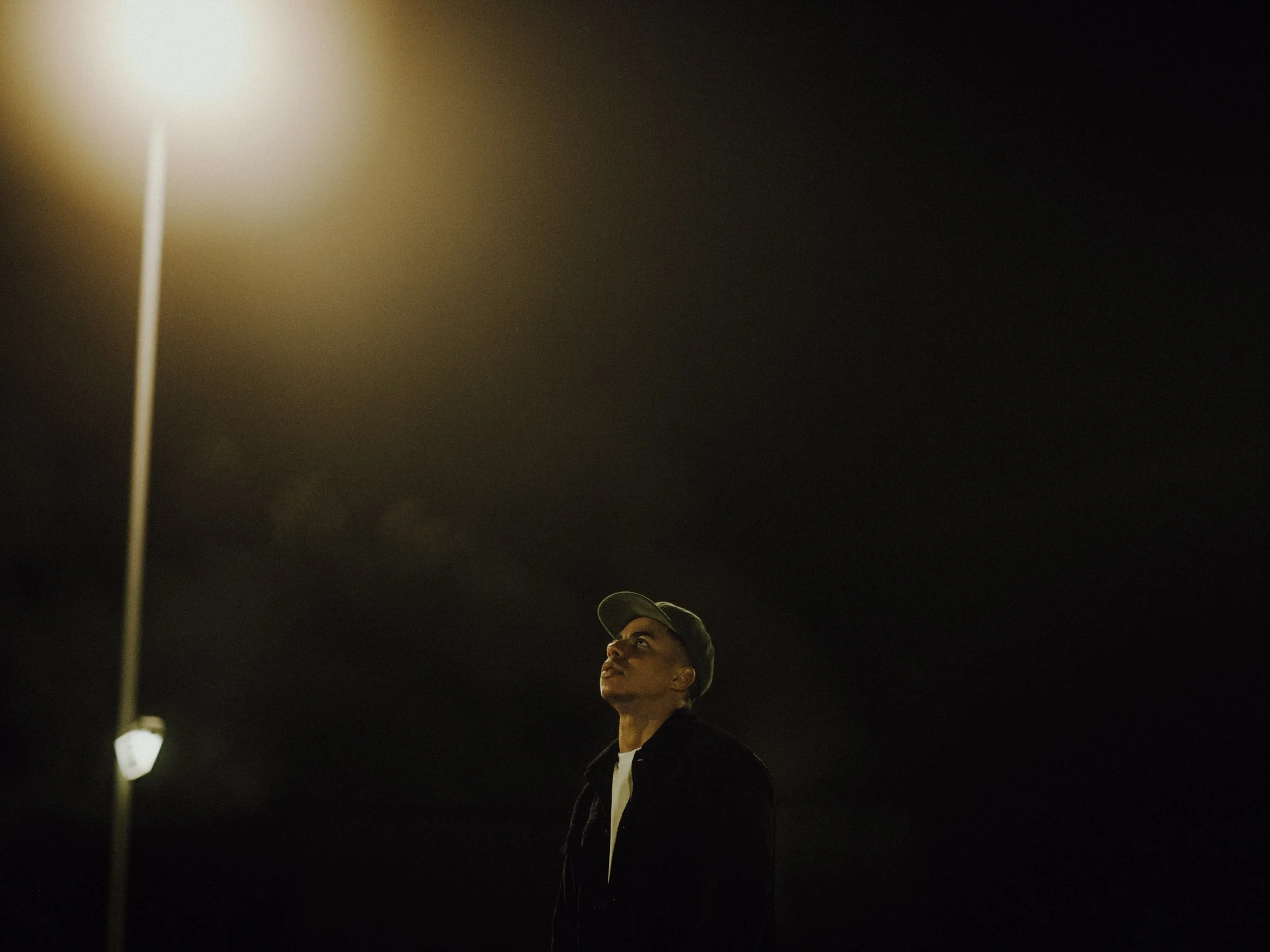 Ein junger Mann steht nachts unter einer Straßenlaterne, schaut nach oben, trägt eine dunkle Jacke und eine Mütze, vor dunklem Himmel.