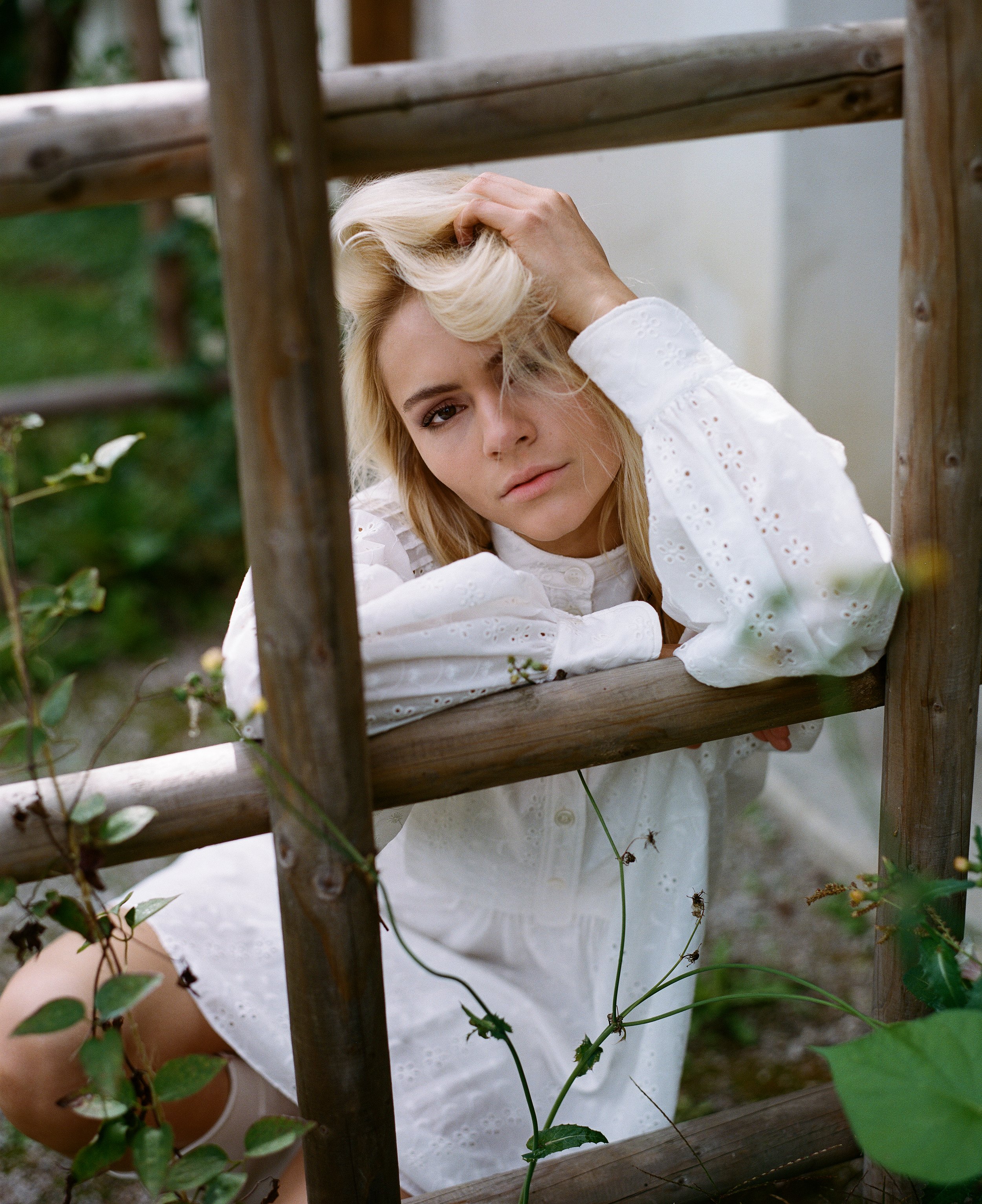 Eine blonde Frau in einem weißen Kleid sitzt hinter einem Holzzaun, stützt den Kopf auf ihren Arm und schaut nach vorne.