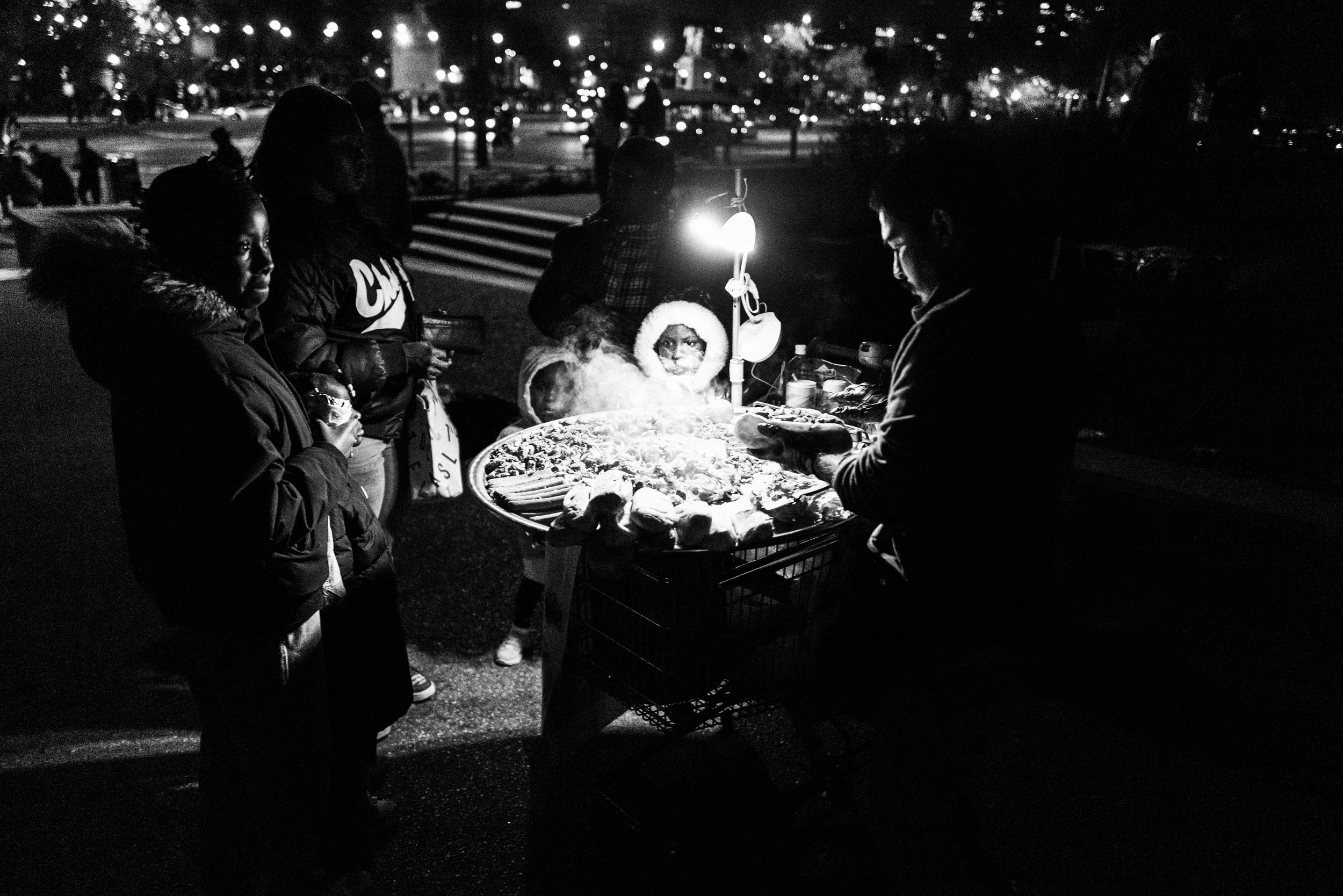 Straßenhändler verkauft Essen an Kunden bei Nacht, Schwarz-Weiß-Fotografie, Menschen vor einem Straßenstand mit Lichter, dunkle Umgebung.