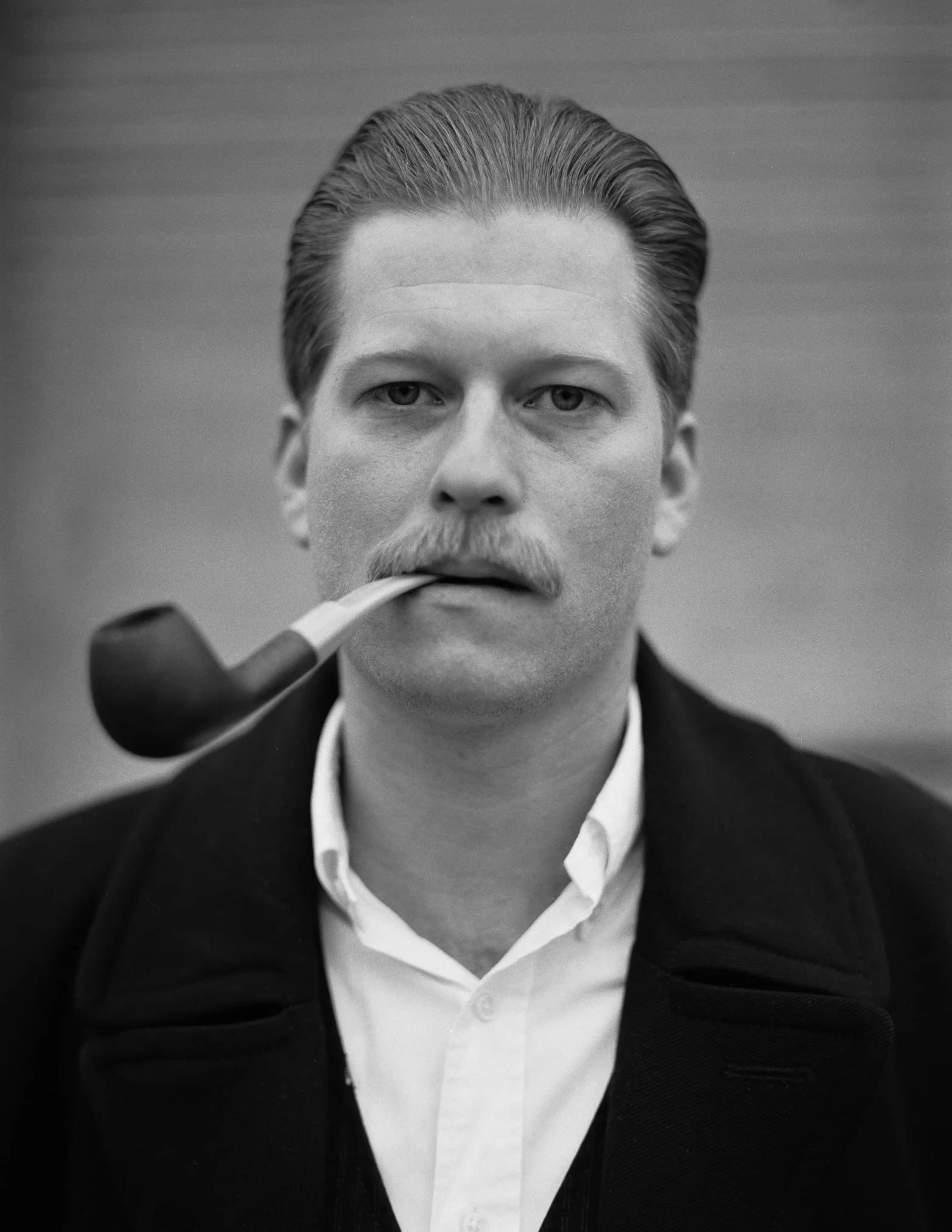 Schwarz-weiß Foto eines Mannes mit Steckbrief, weißem Hemd und dunklem Mantel, der eine Pfeife im Mund hat.